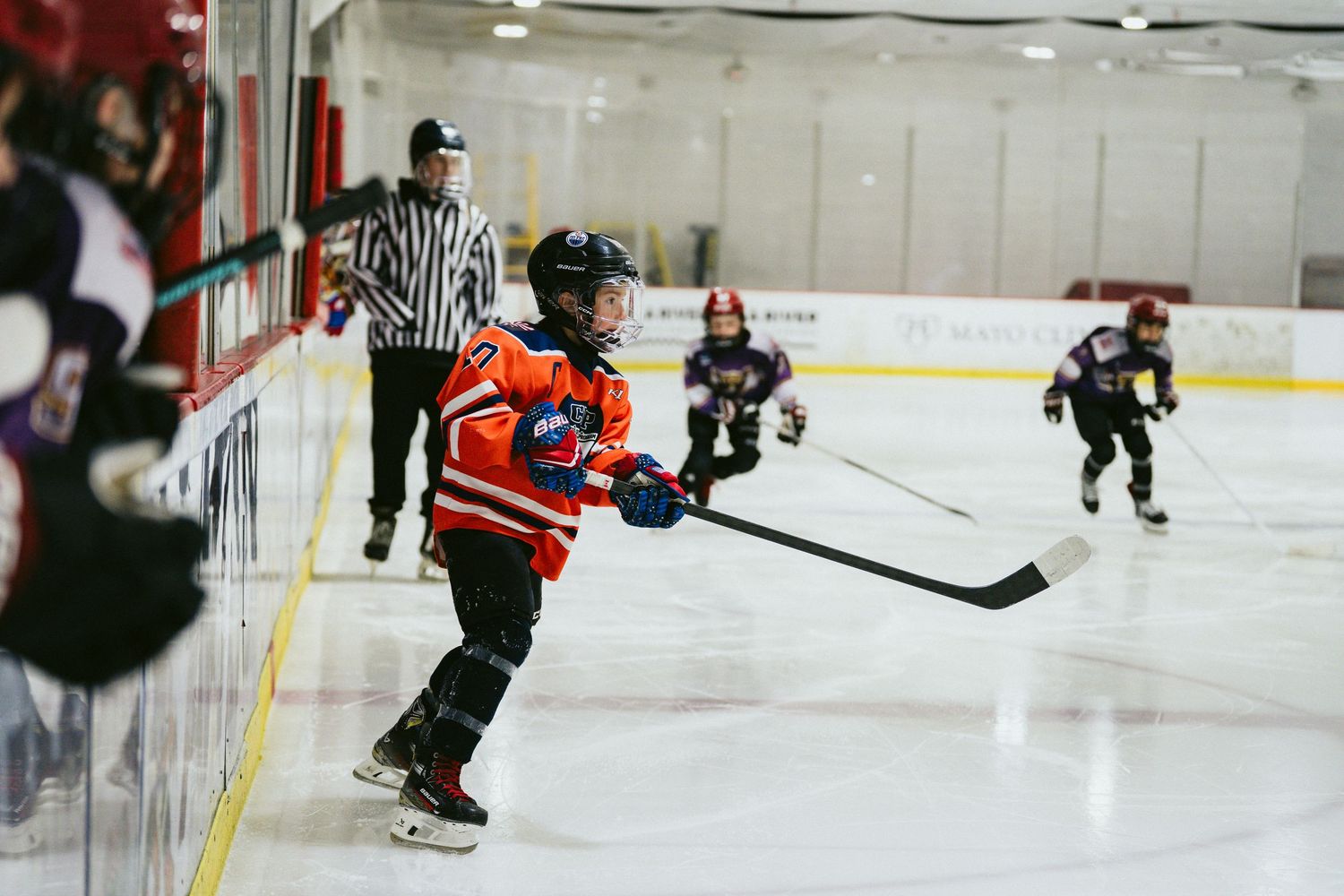 Young hockey player in orange jersey skating near the boards during a youth ice hockey game.