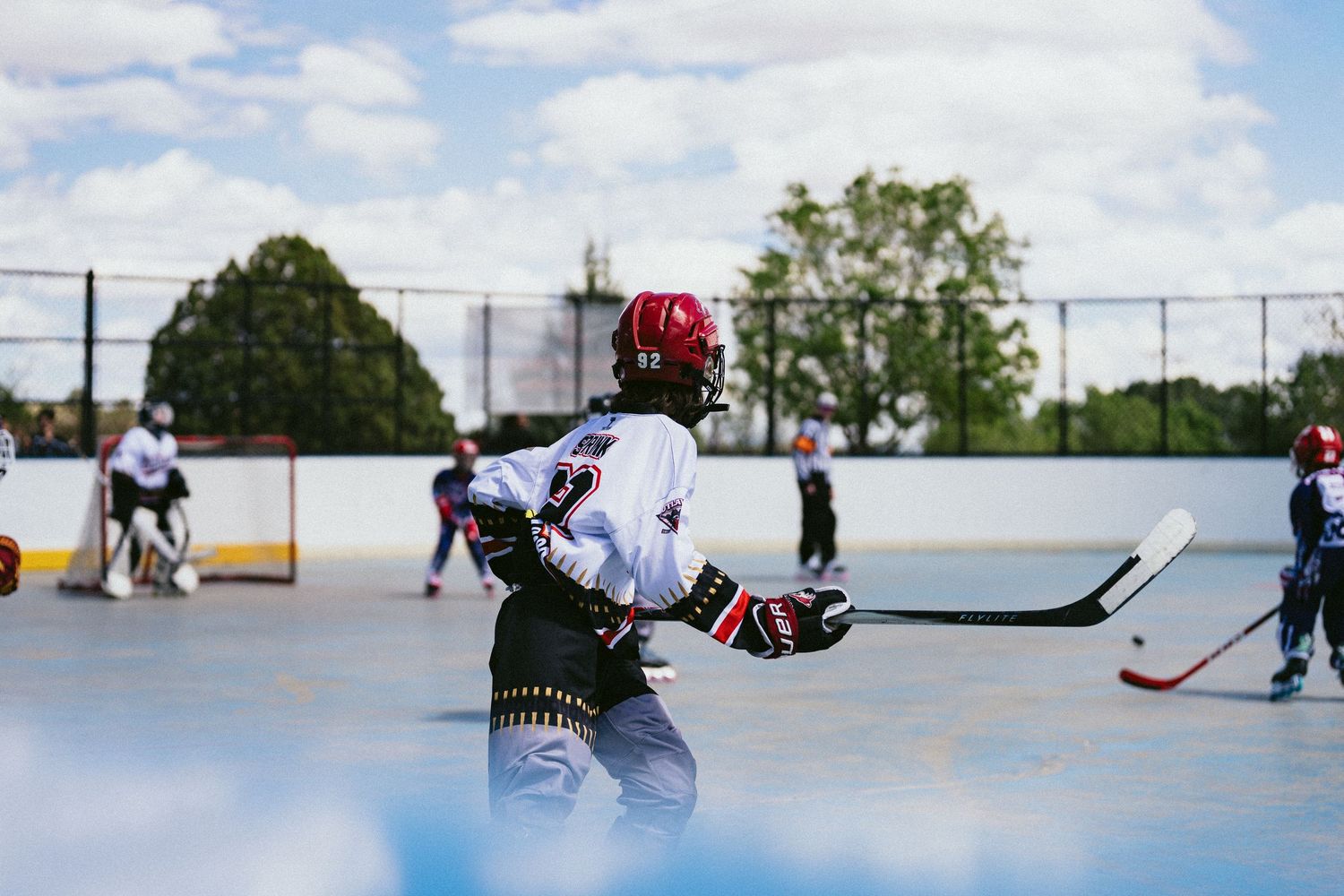 Children playing outdoor roller hockey on a rink under a cloudy sky, wearing helmets and holding hockey sticks.