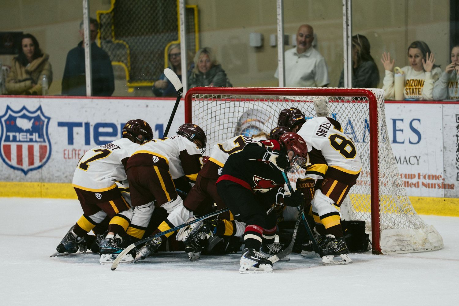 Hockey players pile up near the goal net during an intense game, wearing gold and maroon uniforms on indoor ice.