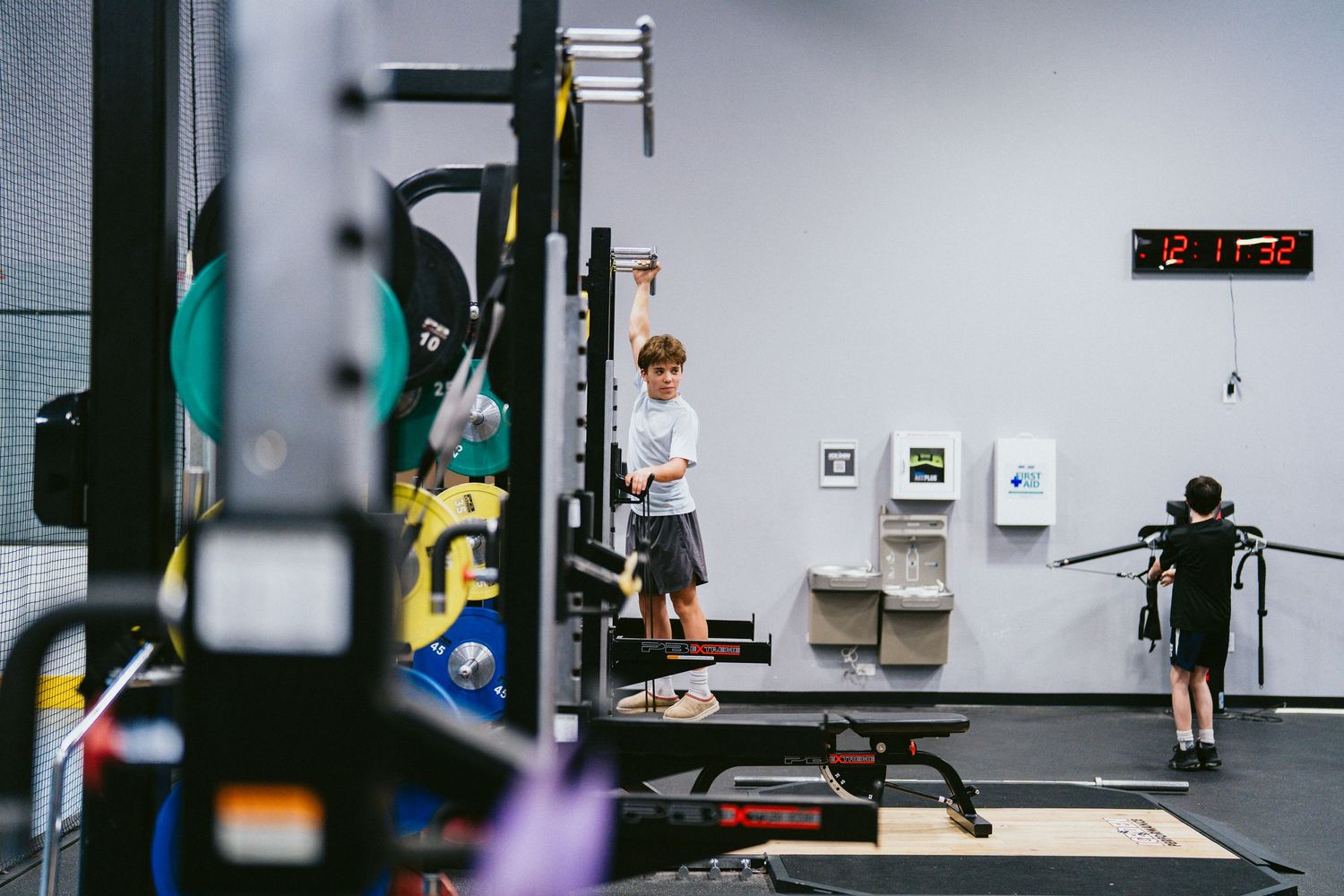 Two children exercising in a modern gym facility with workout equipment and a digital clock on the wall.