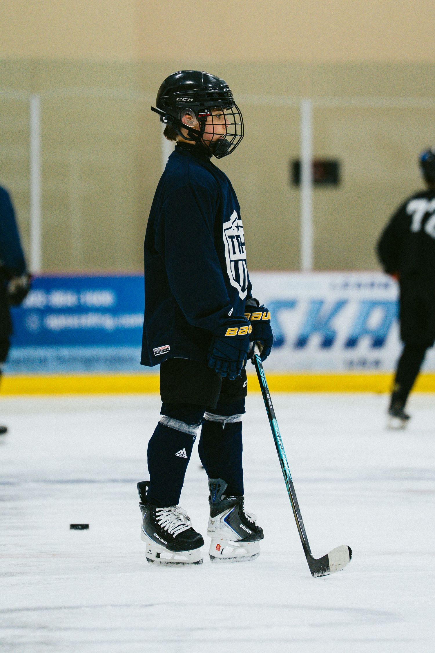 Young hockey player in black uniform and helmet stands on ice rink holding a stick during practice.