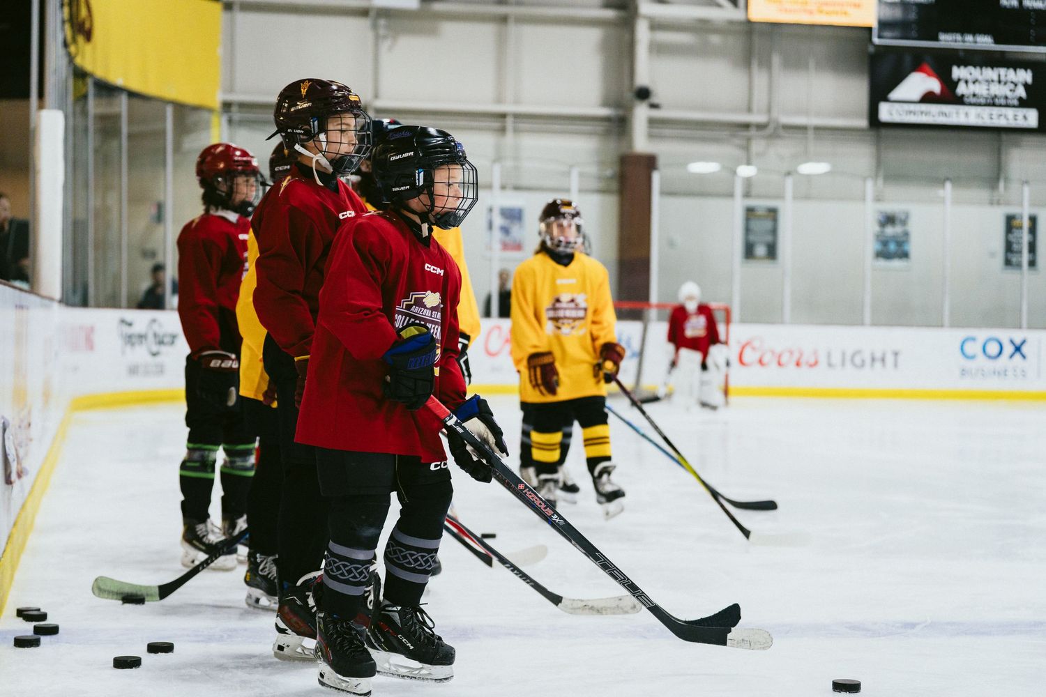 Group of young hockey players in red and yellow jerseys line up with pucks during an ice rink practice.