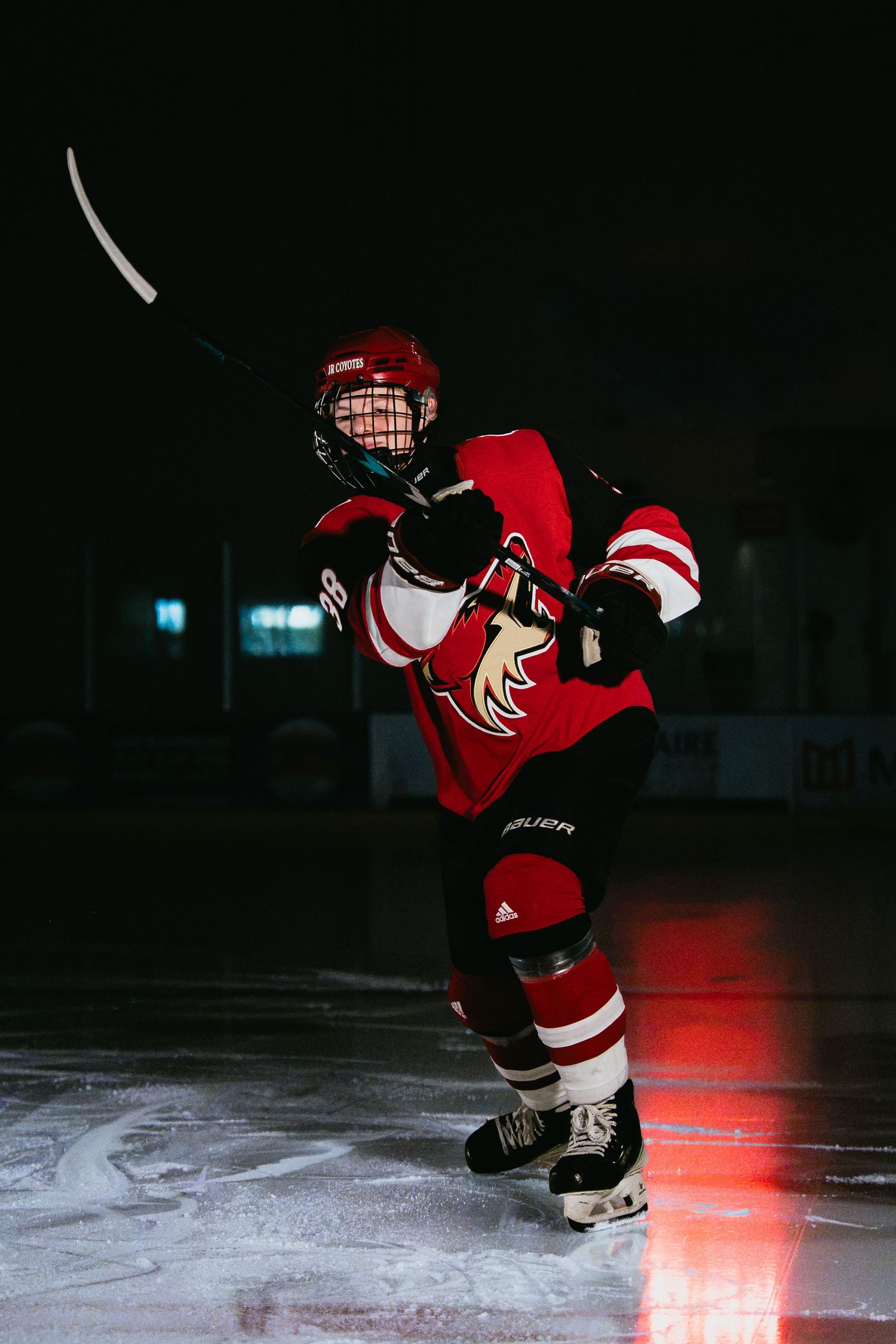 Young hockey player in red jersey and helmet takes a shot on dark ice rink.