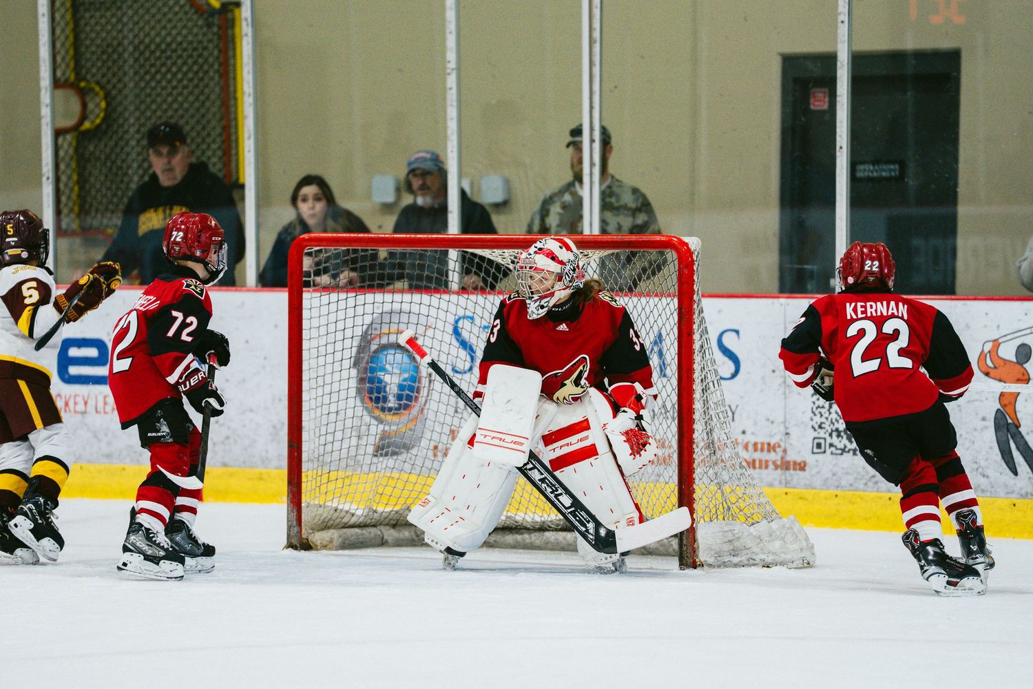 Goalie in red uniform defends the net during intense hockey game action.