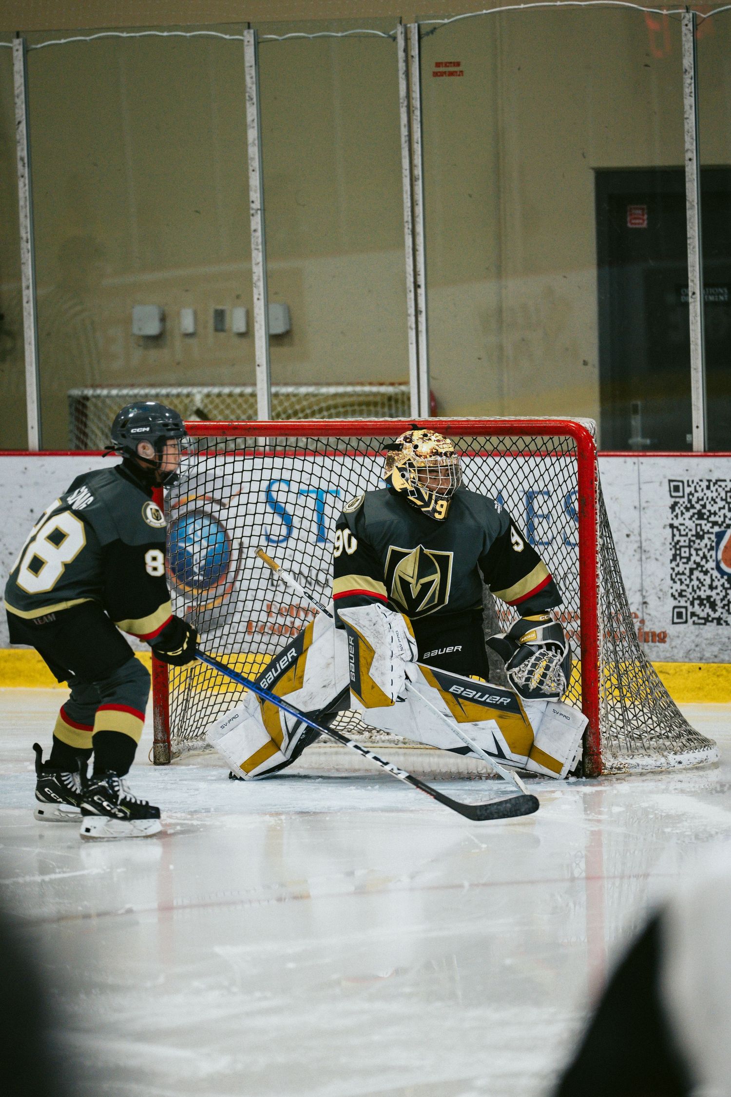Goalie and defenders in black and gold uniforms protect the net during a hockey game.