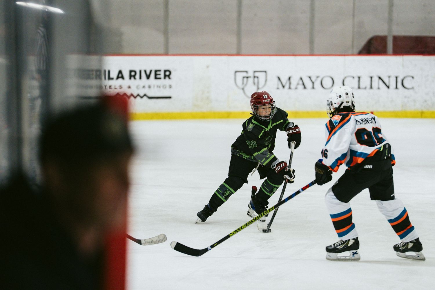 Youth hockey player in green jersey skates with puck during indoor ice hockey game near Mayo Clinic boards.