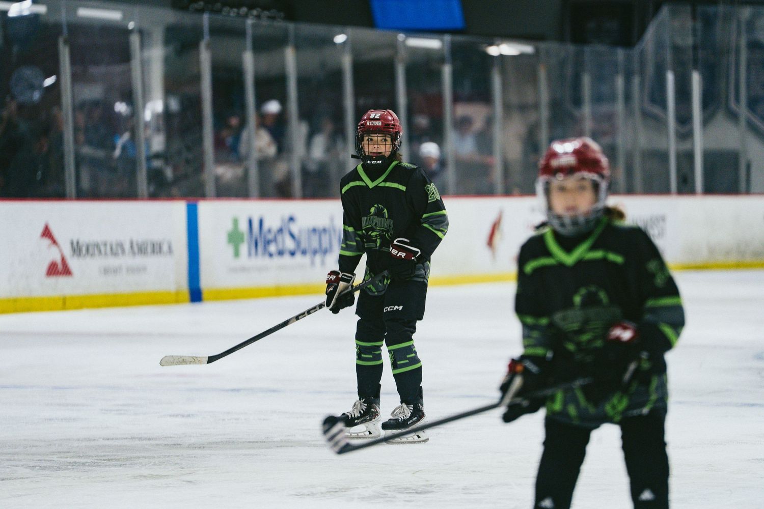 Two young hockey players in black and green uniforms pause during youth ice hockey game on indoor rink.