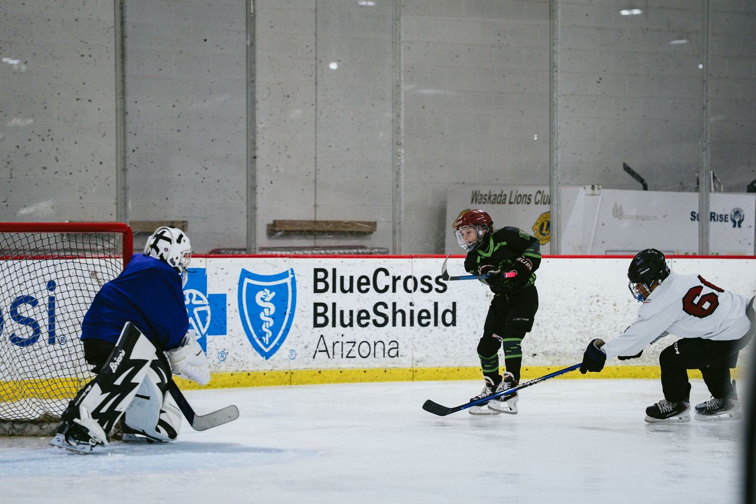Youth hockey player shoots on goal past a blue-uniformed goalie in a BlueCross BlueShield Arizona arena.