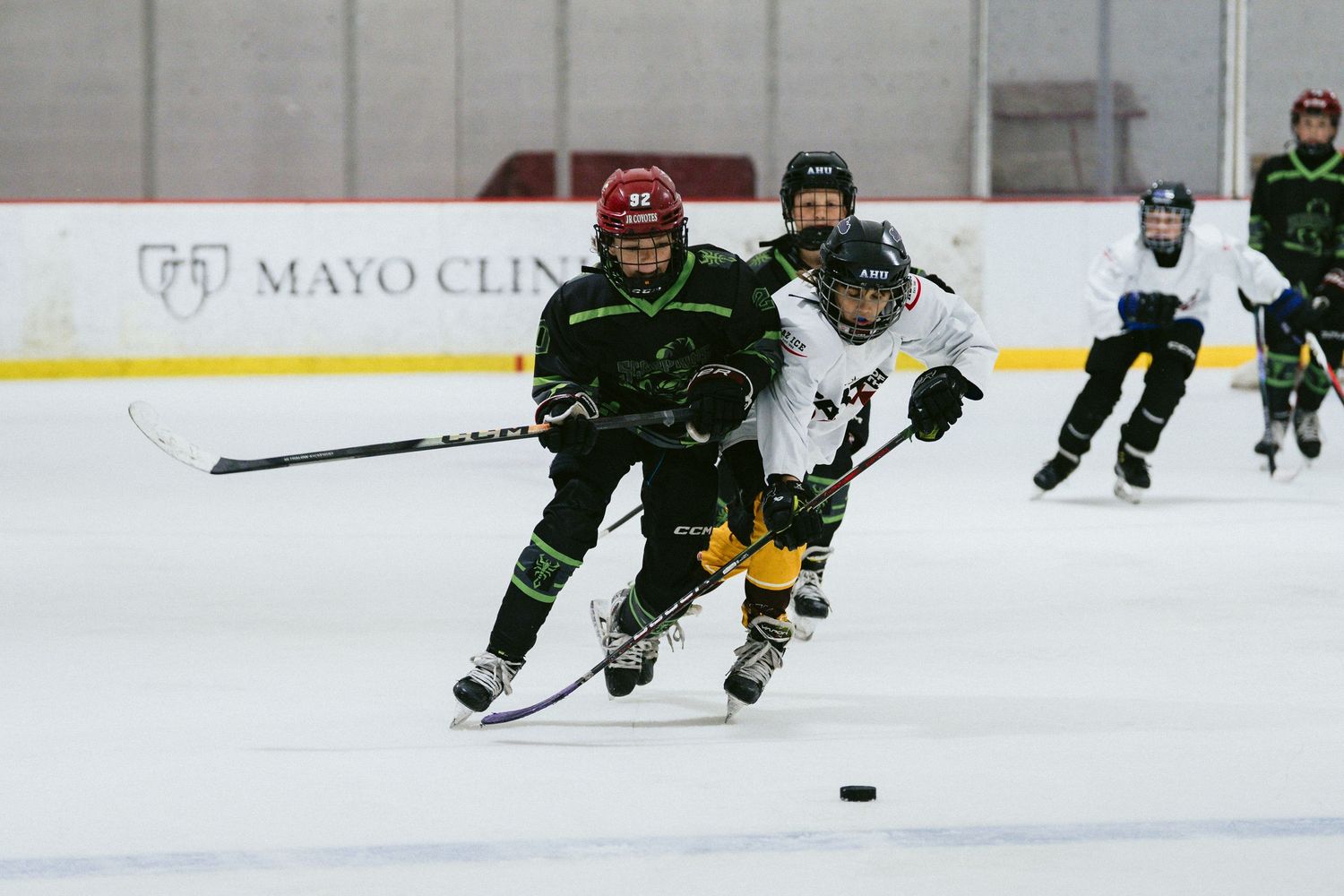 Youth hockey players competing for puck on ice near Mayo Clinic arena boards.