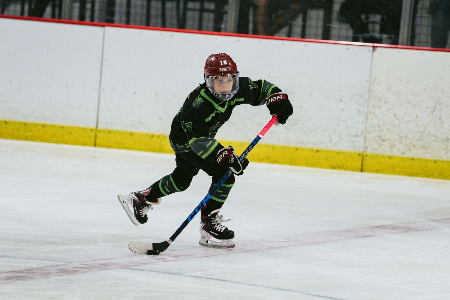 Young hockey player in green uniform skating with stick on indoor ice rink during a game.