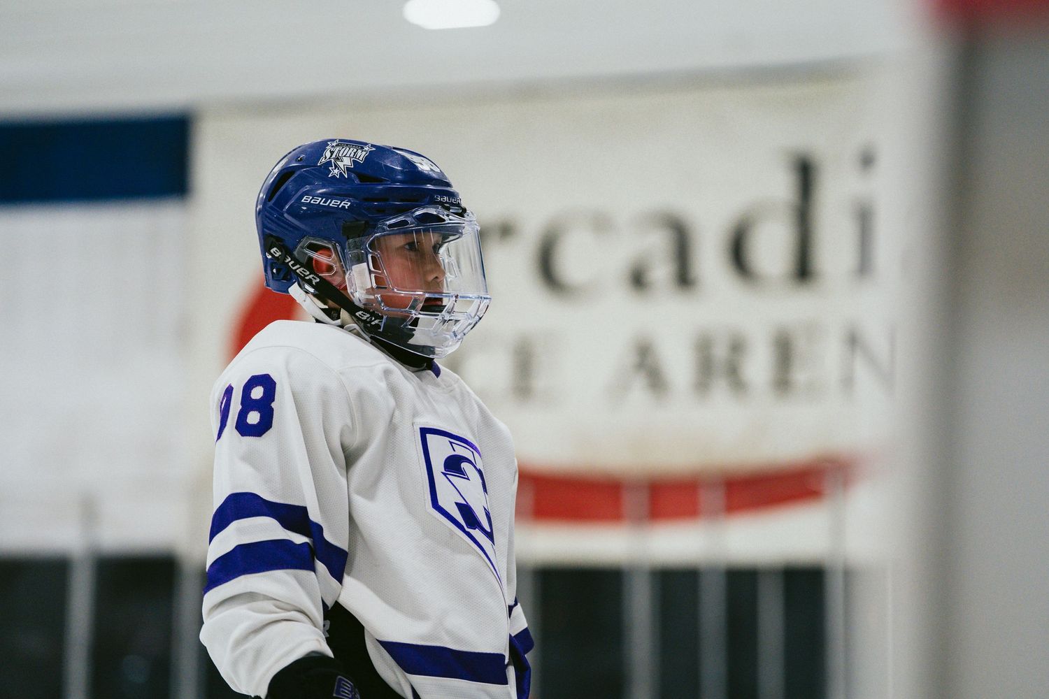Hockey player in white jersey and blue helmet stands alert on ice rink.