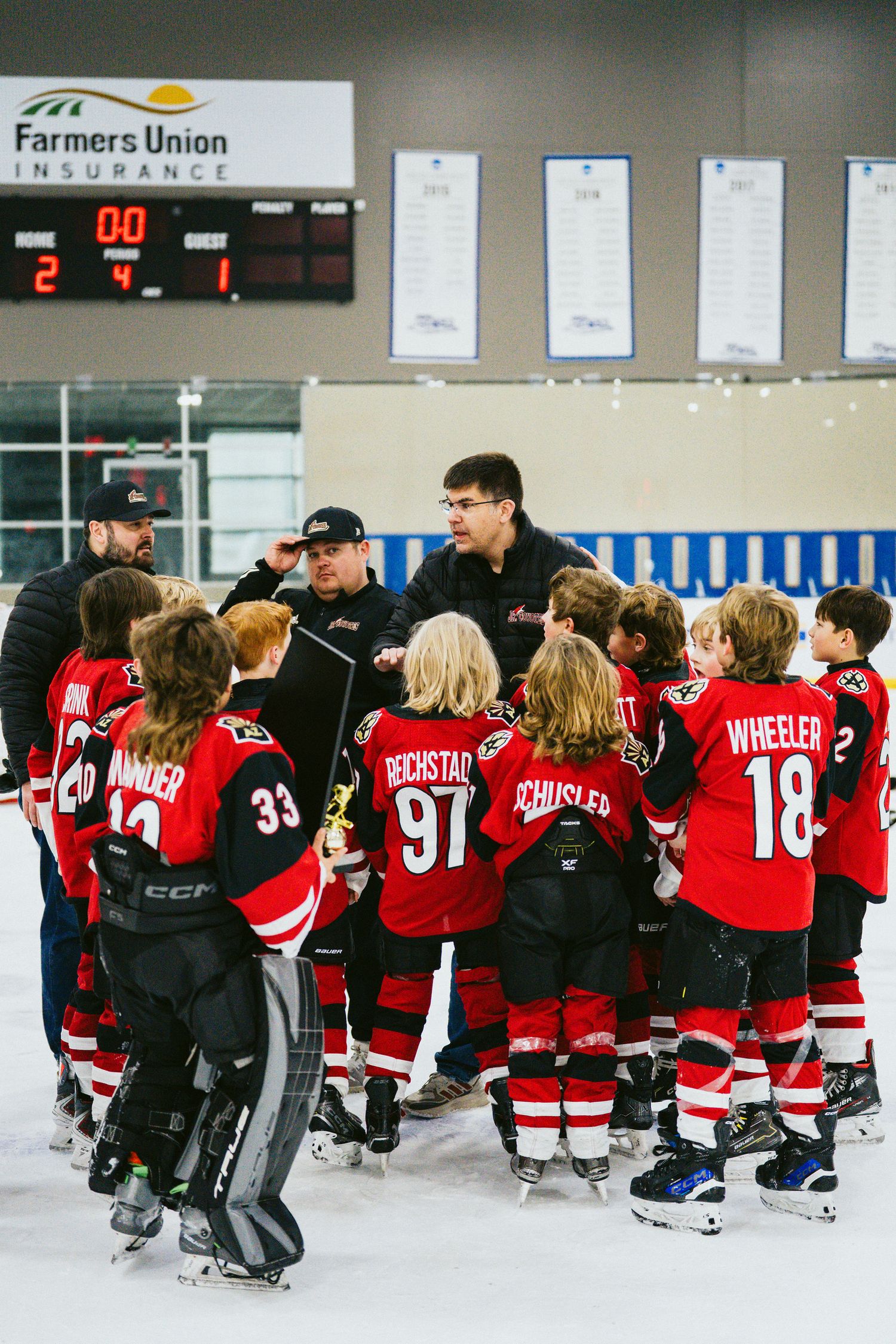 Youth hockey players in red jerseys gathered on ice rink under scoreboard at Farmers Union arena.