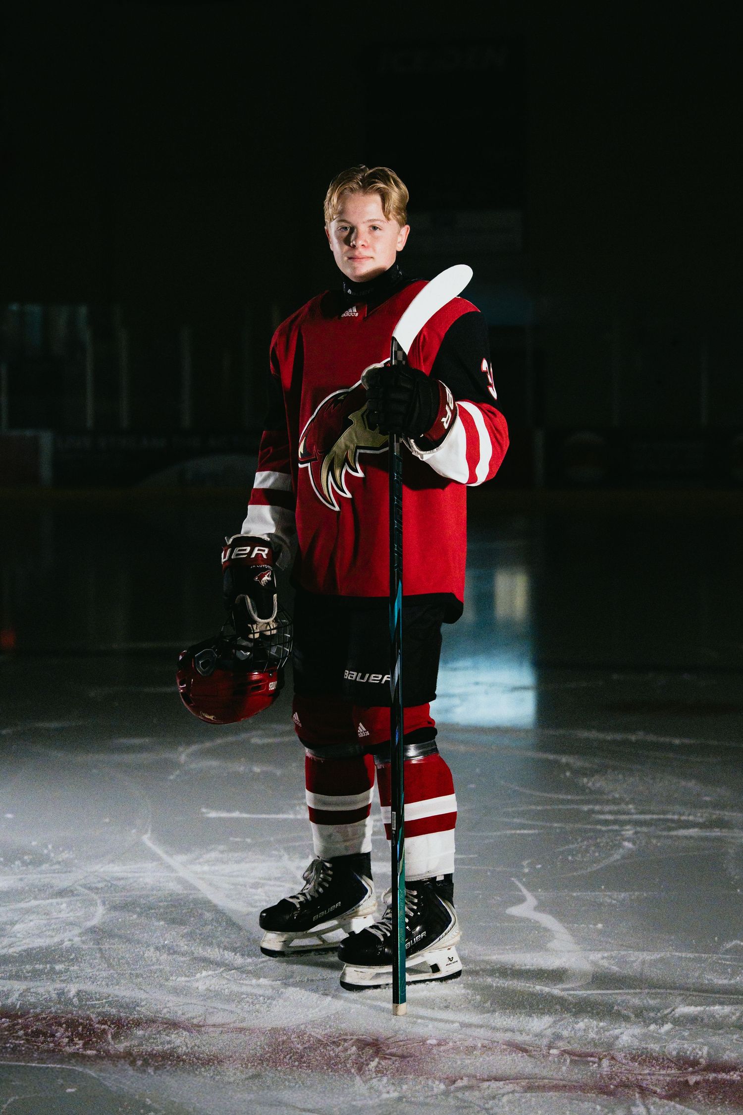 Young hockey player in red uniform holding stick and helmet on ice rink in dramatic dark lighting.