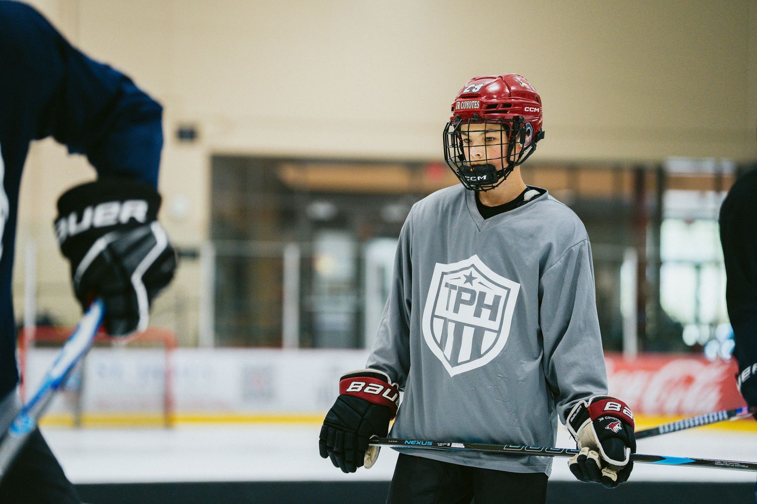 Young hockey player in red helmet and TPH gray jersey stands attentively during ice hockey practice.