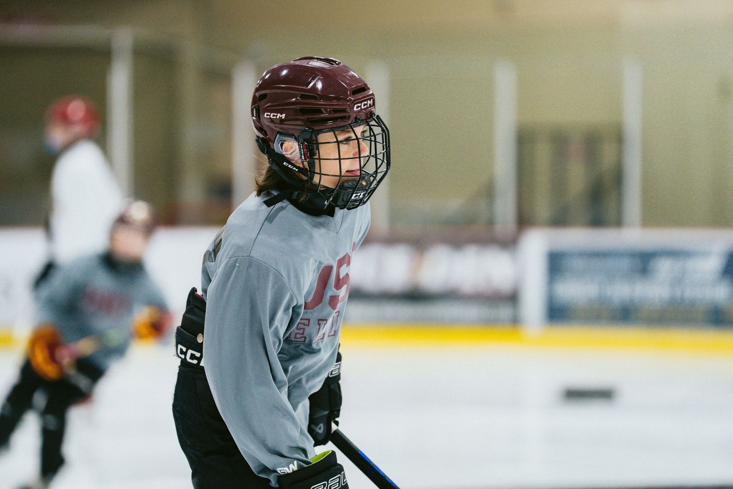 Young hockey player in maroon helmet and gray jersey focuses during ice hockey practice at an indoor rink.