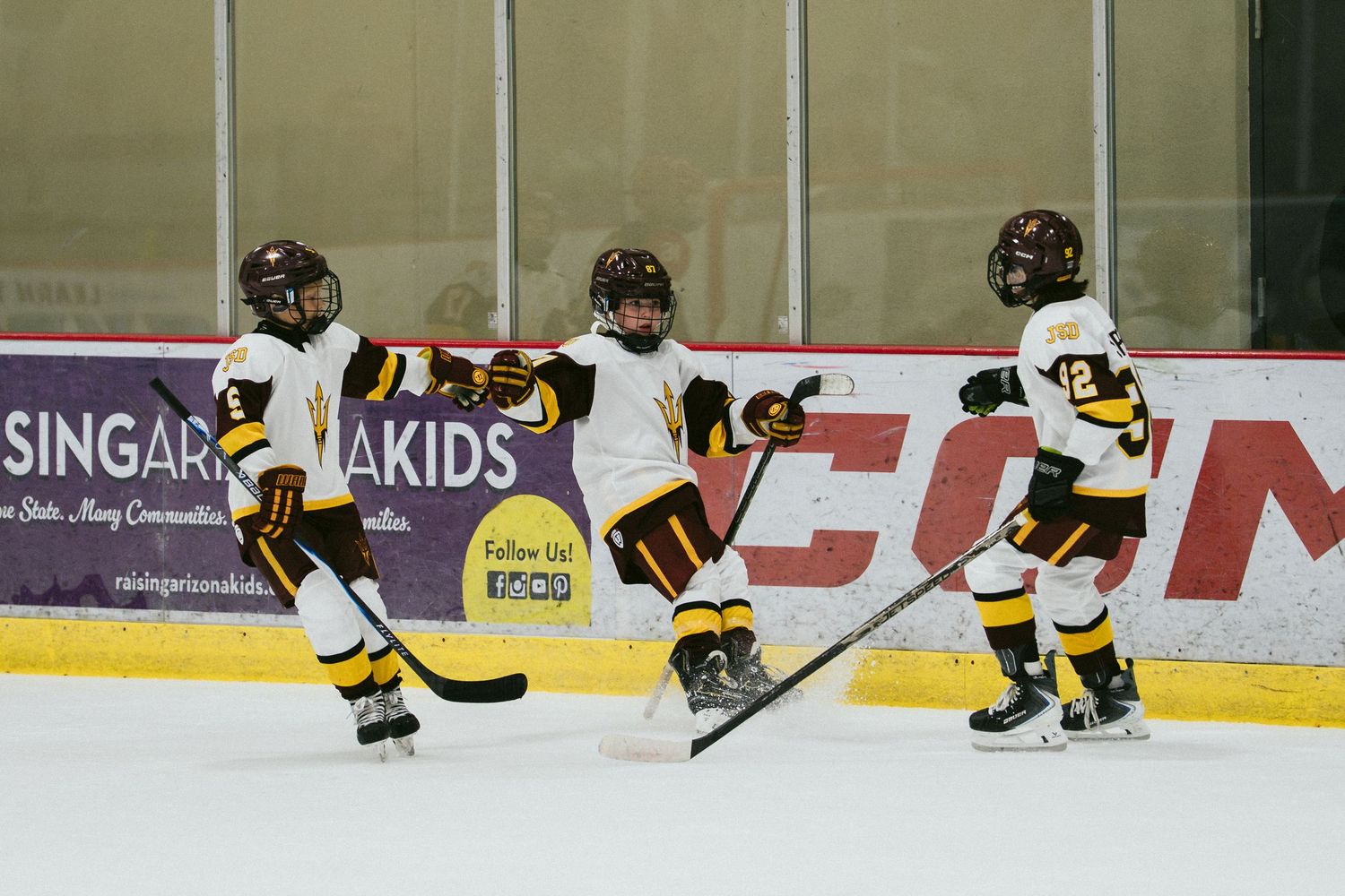 Three young children in hockey uniforms skating on an indoor ice rink during a youth hockey game.