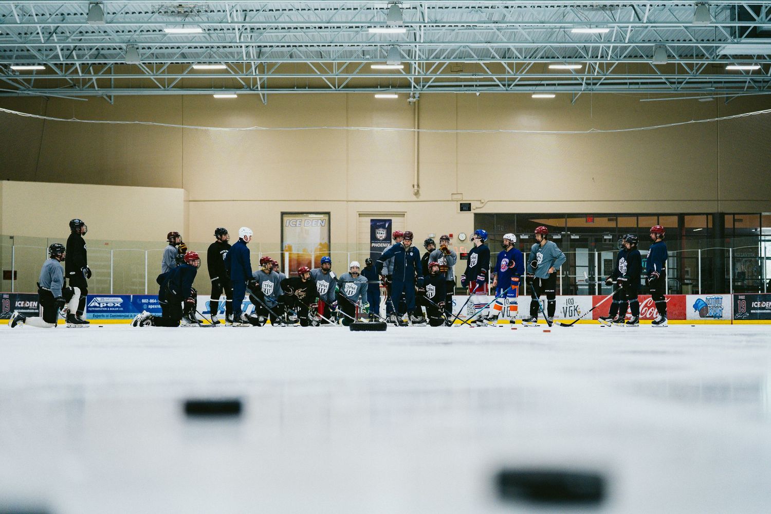 Hockey players and coaches gather on an indoor ice rink during a practice session, with pucks scattered on the ice.
