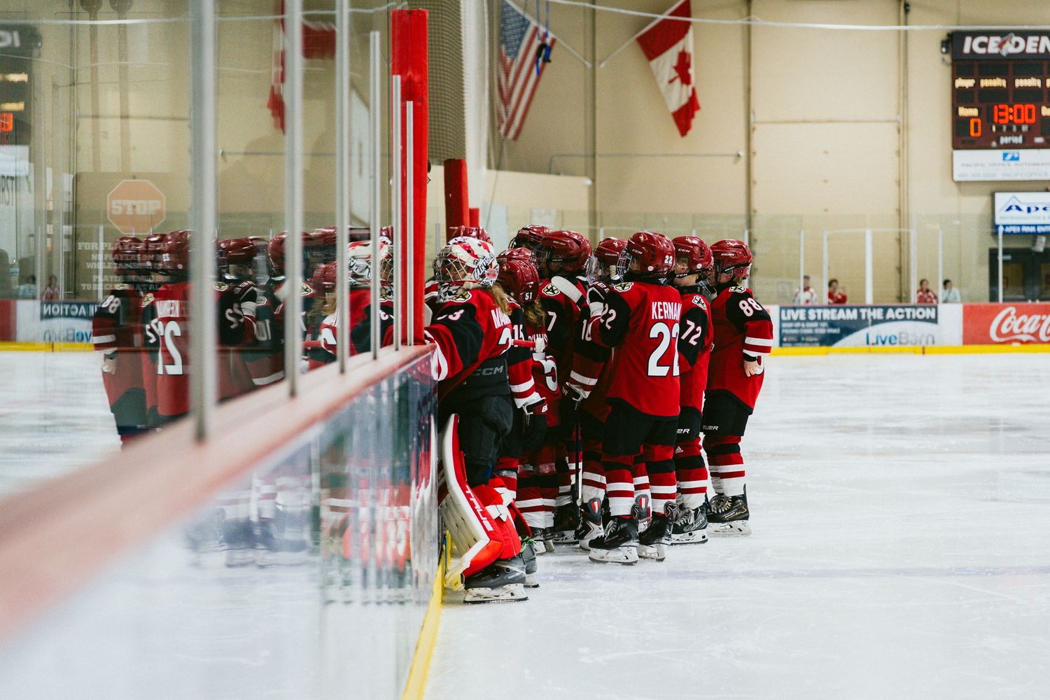 Hockey players in red and black uniforms sit on benches during a game in an indoor ice rink.