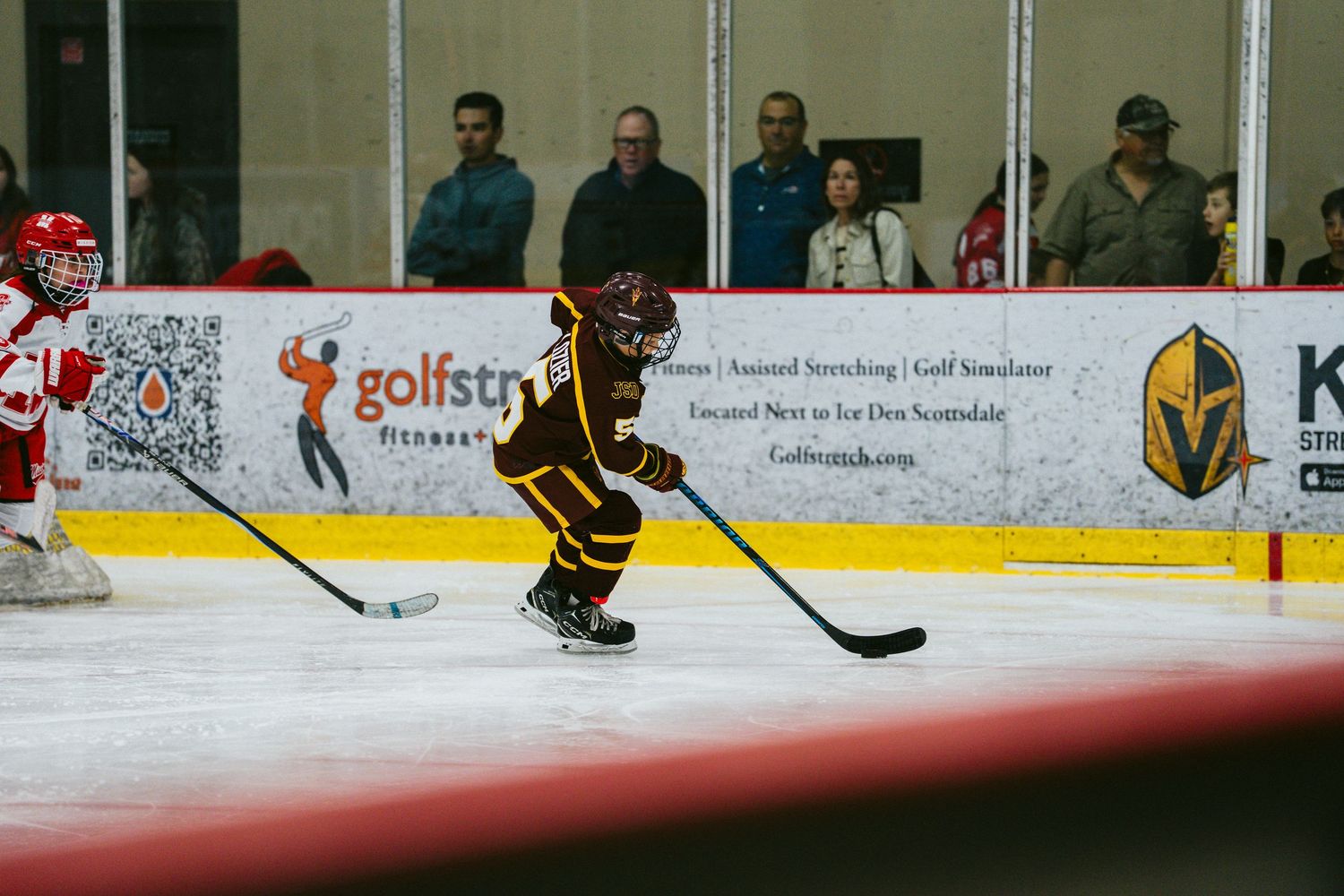 Youth hockey players in black and gold jerseys compete during an intense ice hockey match at an indoor rink.