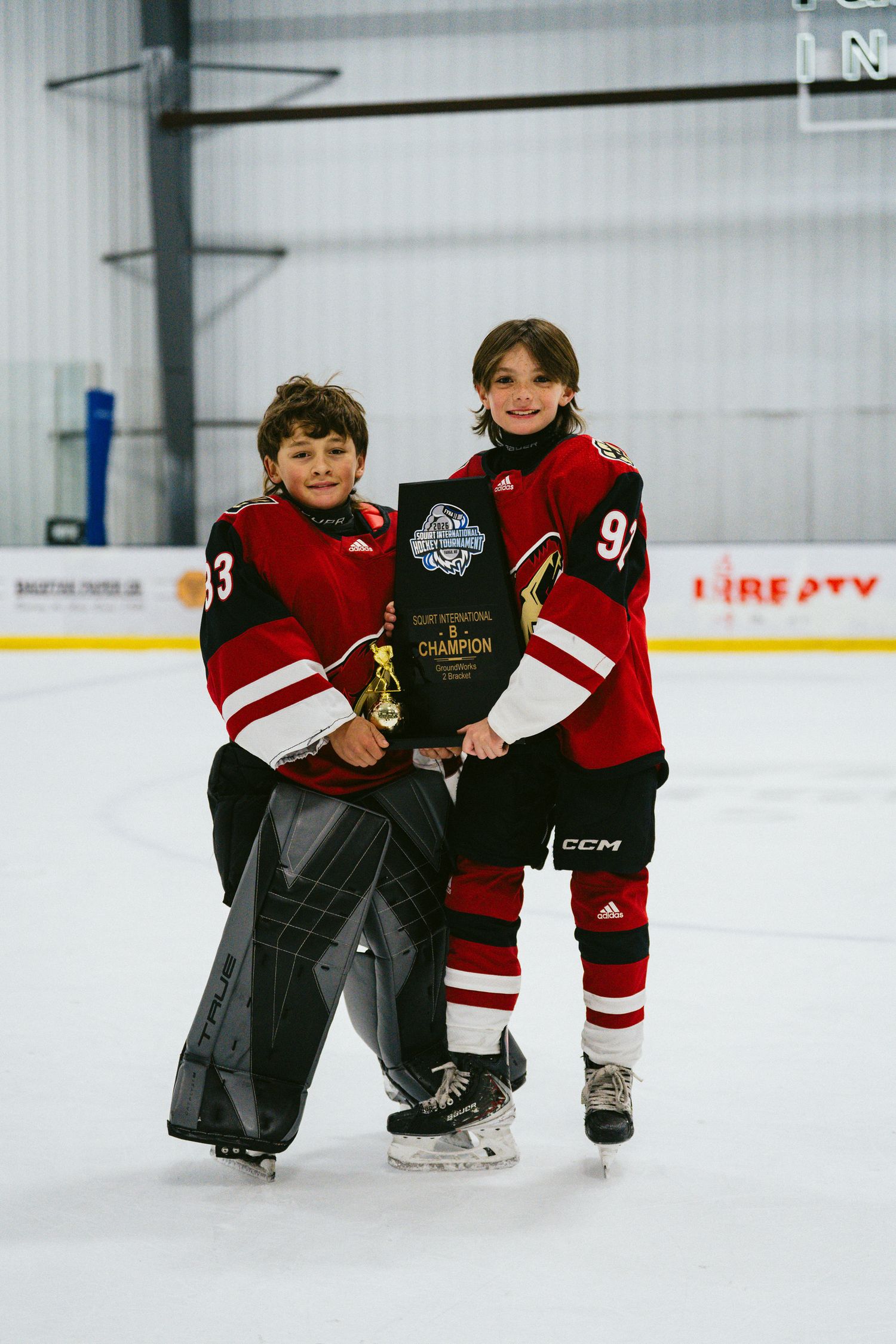 Young hockey players in red and black jerseys sitting together on ice rink.