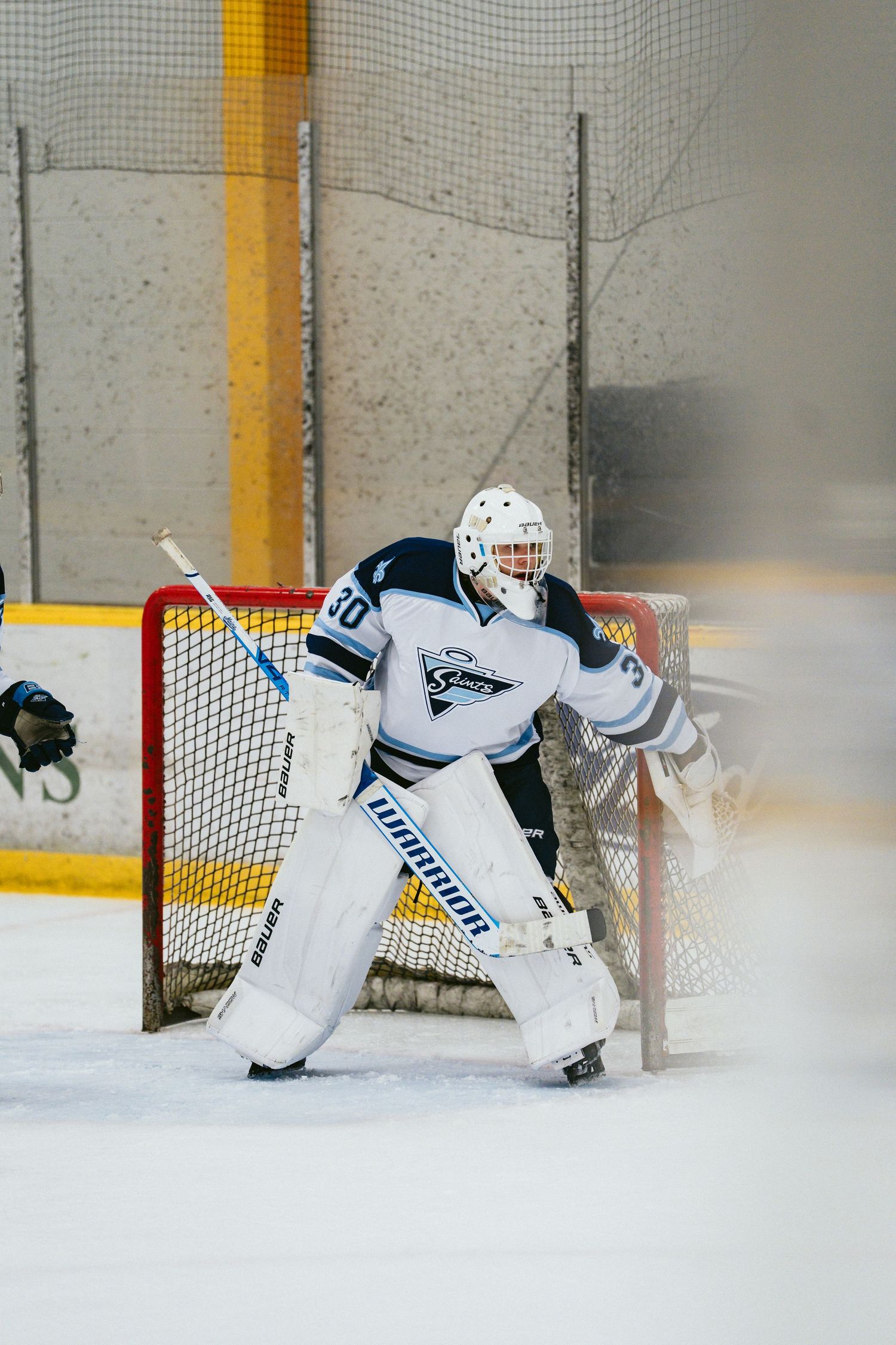 Hockey goalie in white Warrior gear defends the net during an indoor ice hockey game.