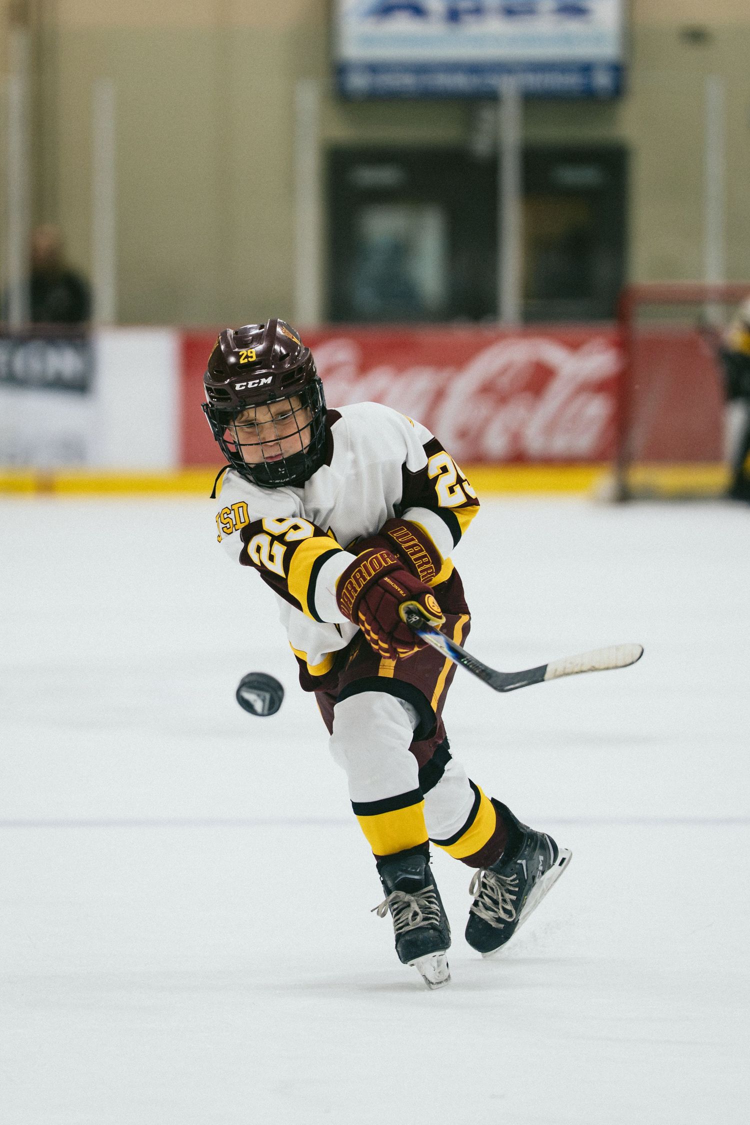 Young ice hockey player in maroon and gold uniform shooting a puck during a youth league game at an indoor rink.