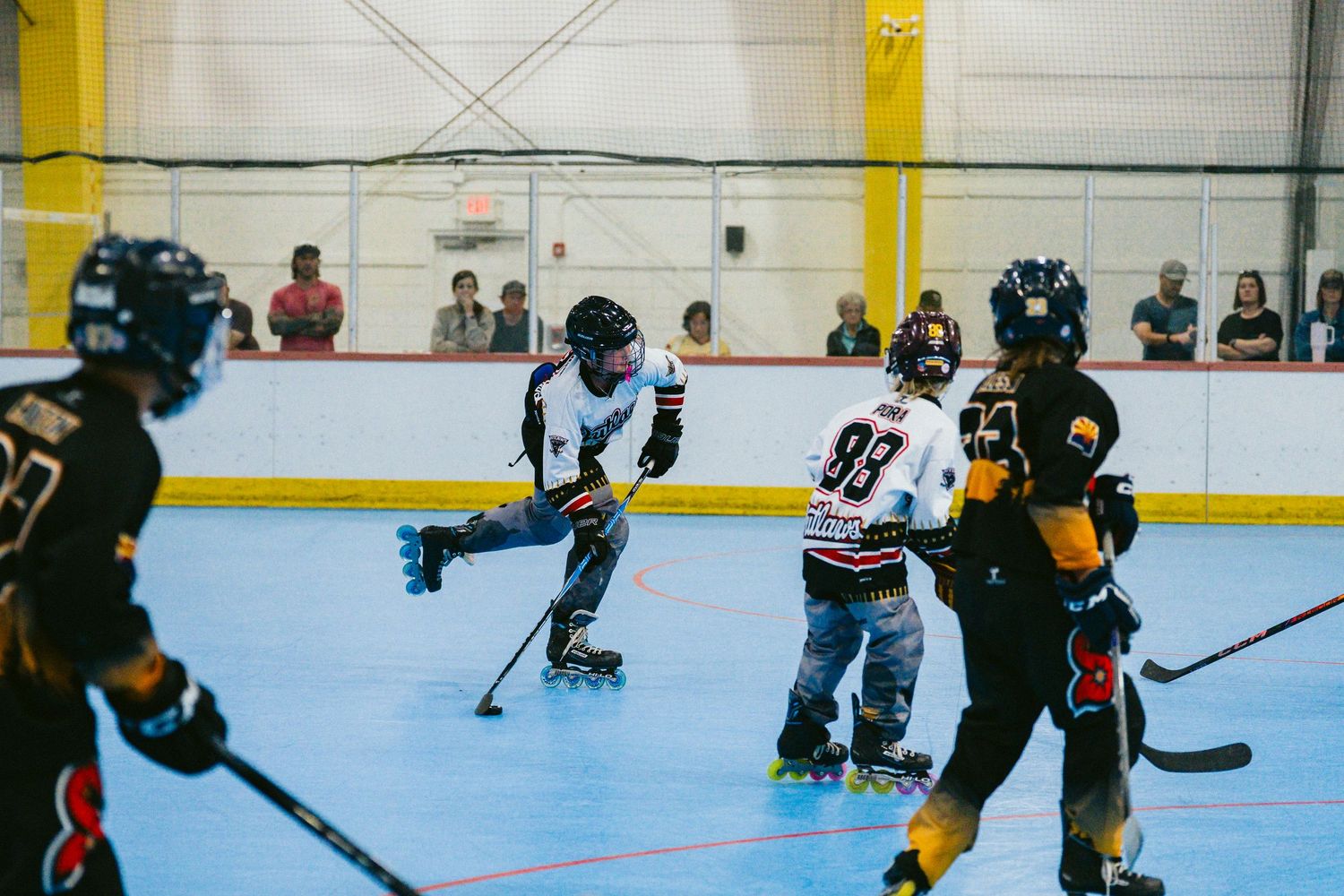 Indoor roller hockey game with players in black and white uniforms competing on a rink with spectators watching.