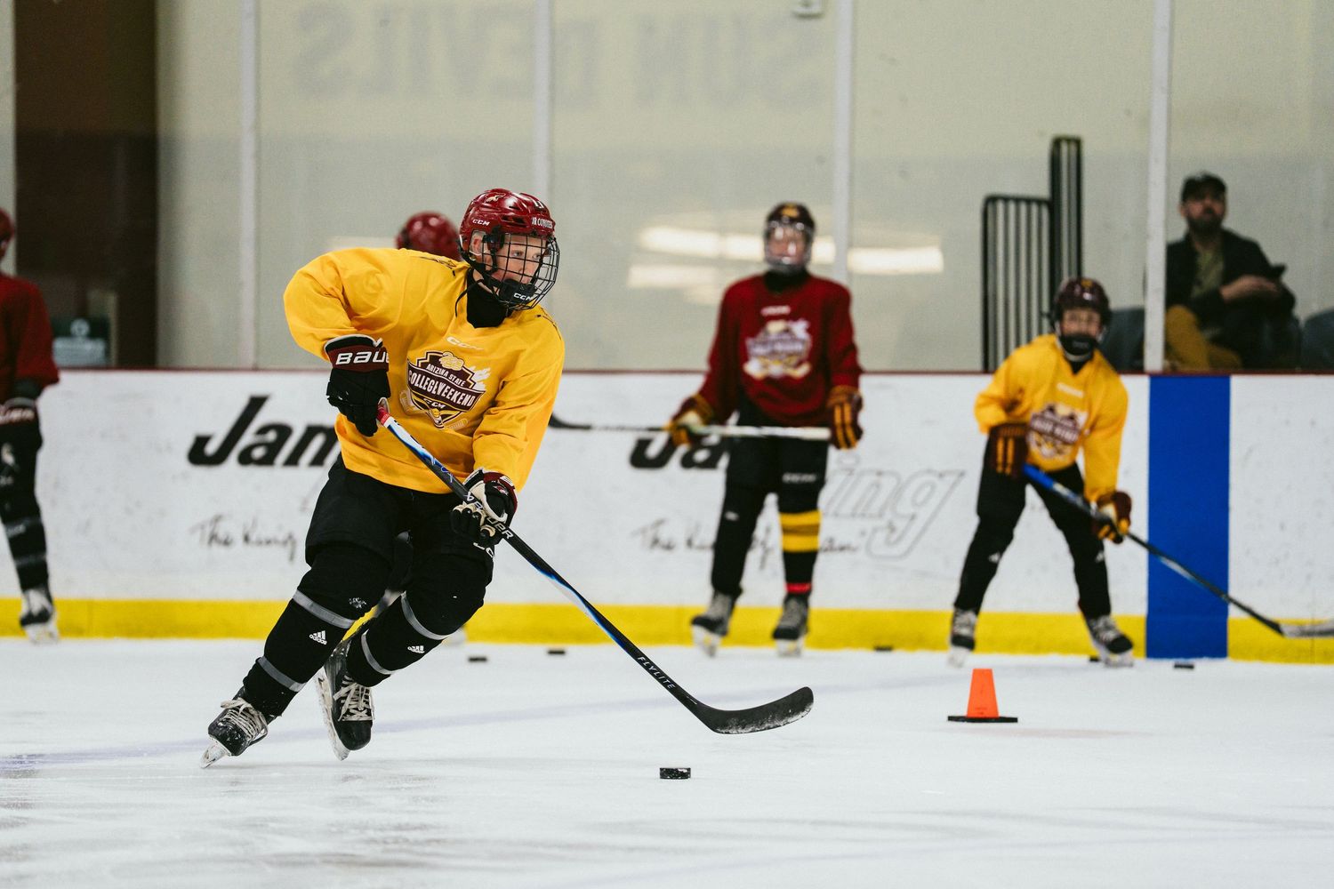 Youth hockey player in yellow jersey skates with puck during indoor ice rink drill with teammates watching.