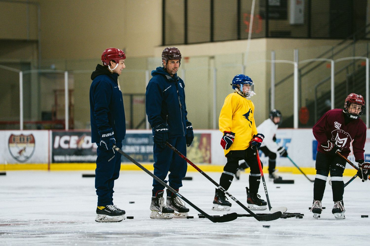 Two coaches instructing young hockey players during ice skating practice at an indoor hockey rink.
