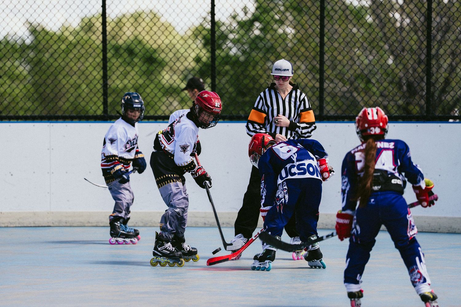 Kids playing roller hockey with referee during outdoor inline hockey game on a rink.