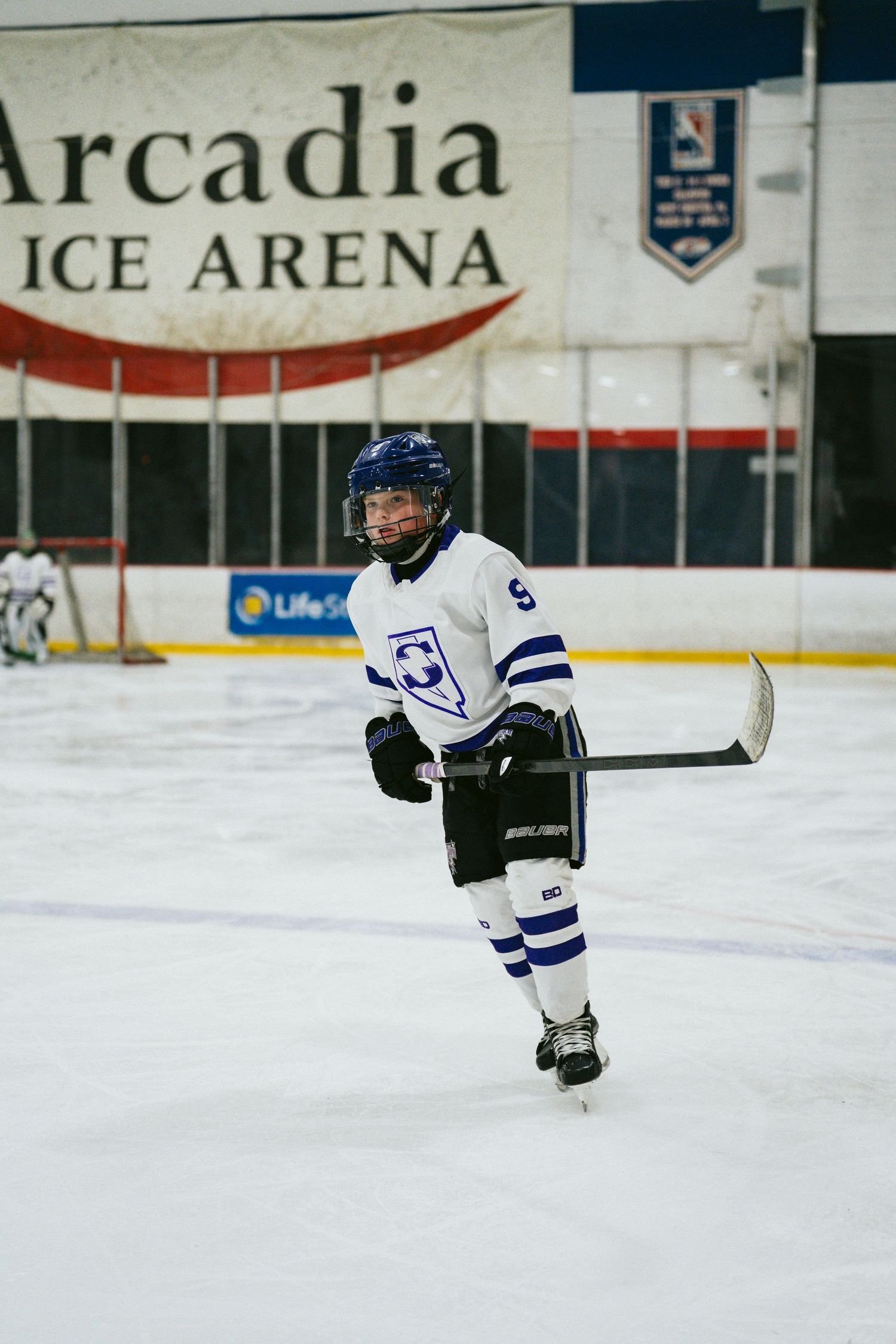 Young hockey player gliding across ice rink in white and blue team uniform.