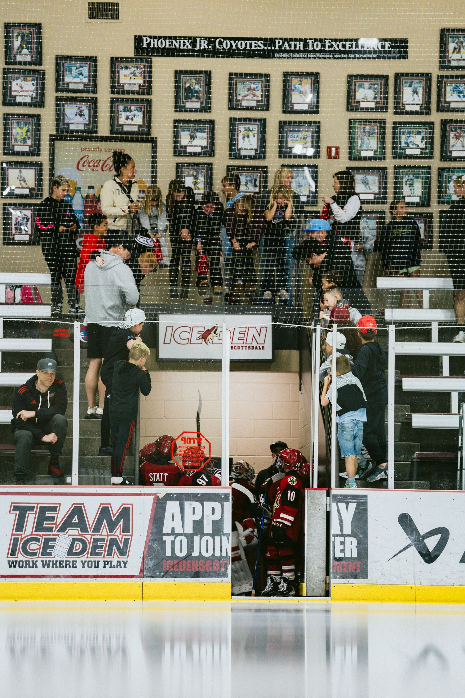 Hockey spectators watch from behind glass barriers at an indoor ice rink with advertisements visible.