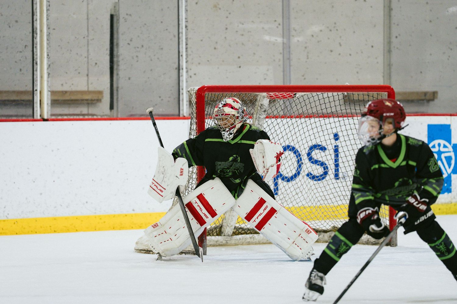 Youth hockey goalie in green and white gear guards the net during an indoor ice hockey game.