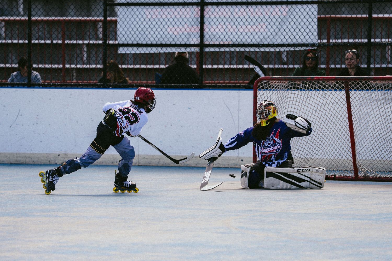 Young roller hockey player attempts a shot on goal against a diving goalie in an outdoor rink game.