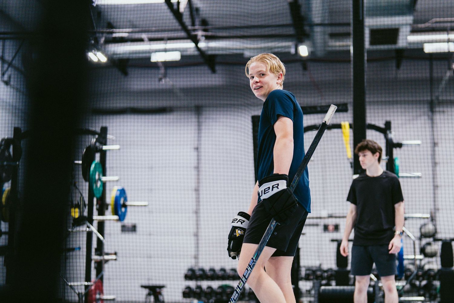 Young hockey player holding a stick in an indoor training facility gym with weights and equipment in background.