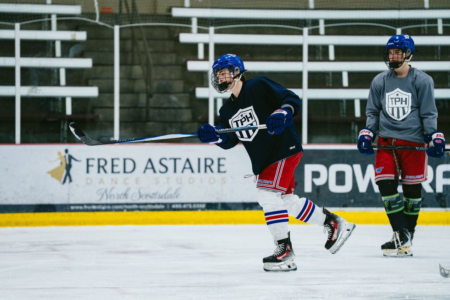 Hockey player in Rangers-style uniform skates during practice with teammate watching near Fred Astaire advertisement.