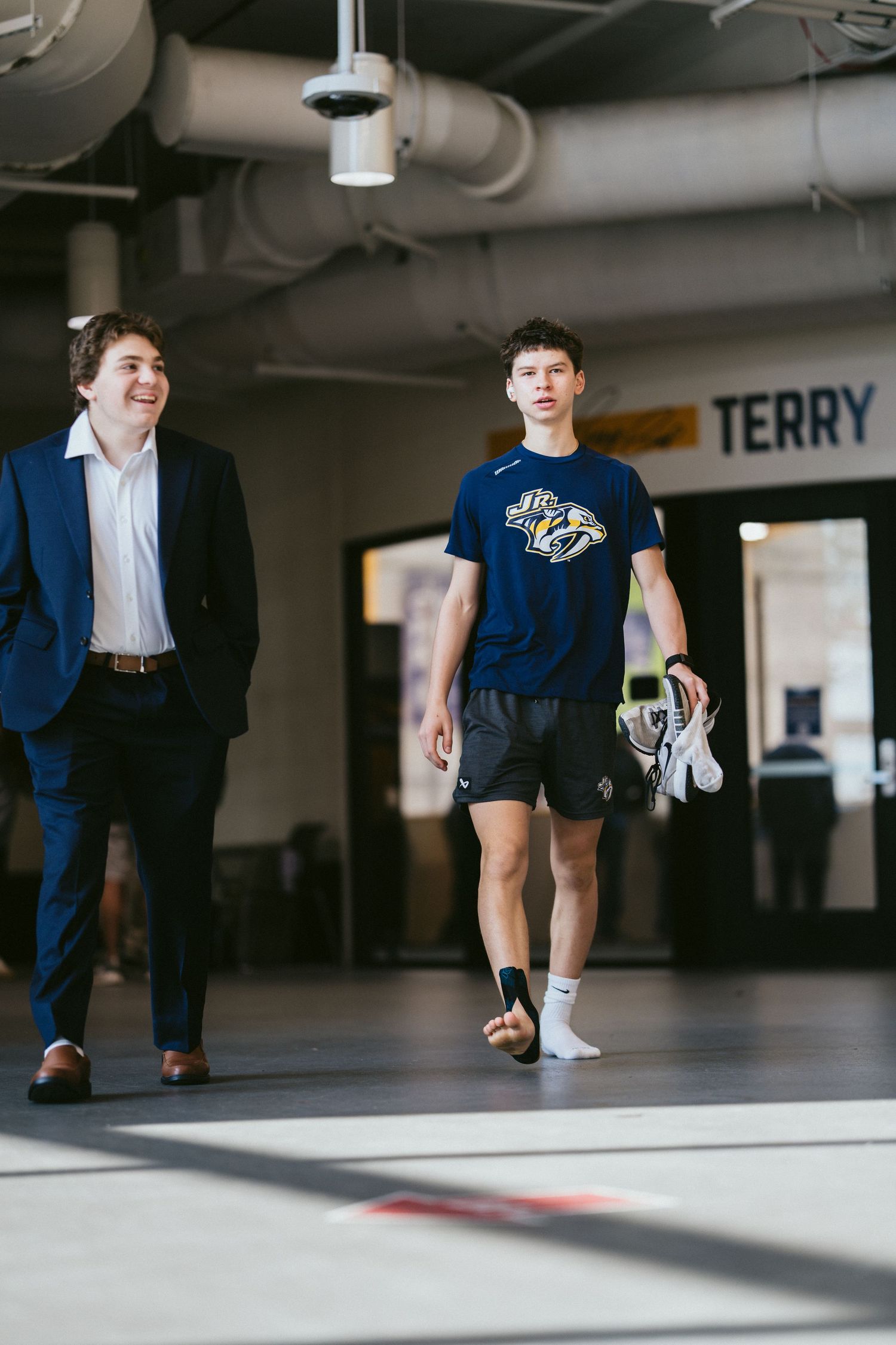 Two young men walk through an indoor arena corridor, one in a suit, one in Nashville Predators gear.