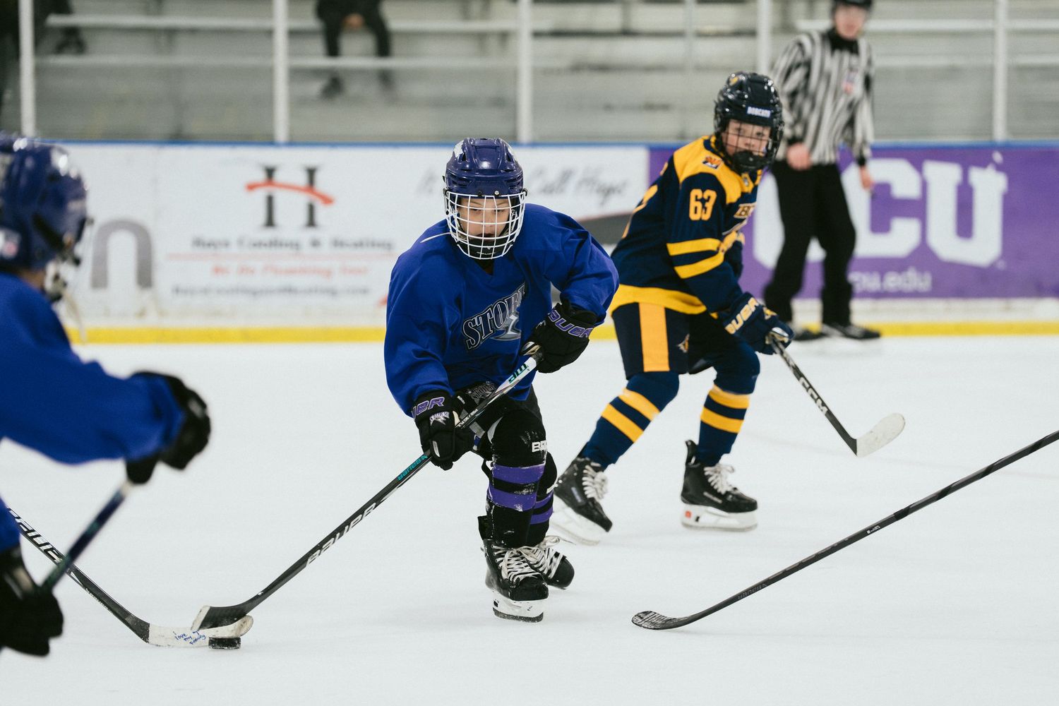 Young hockey players compete on indoor ice rink, player in blue jersey controls puck against opponent in gold uniform.