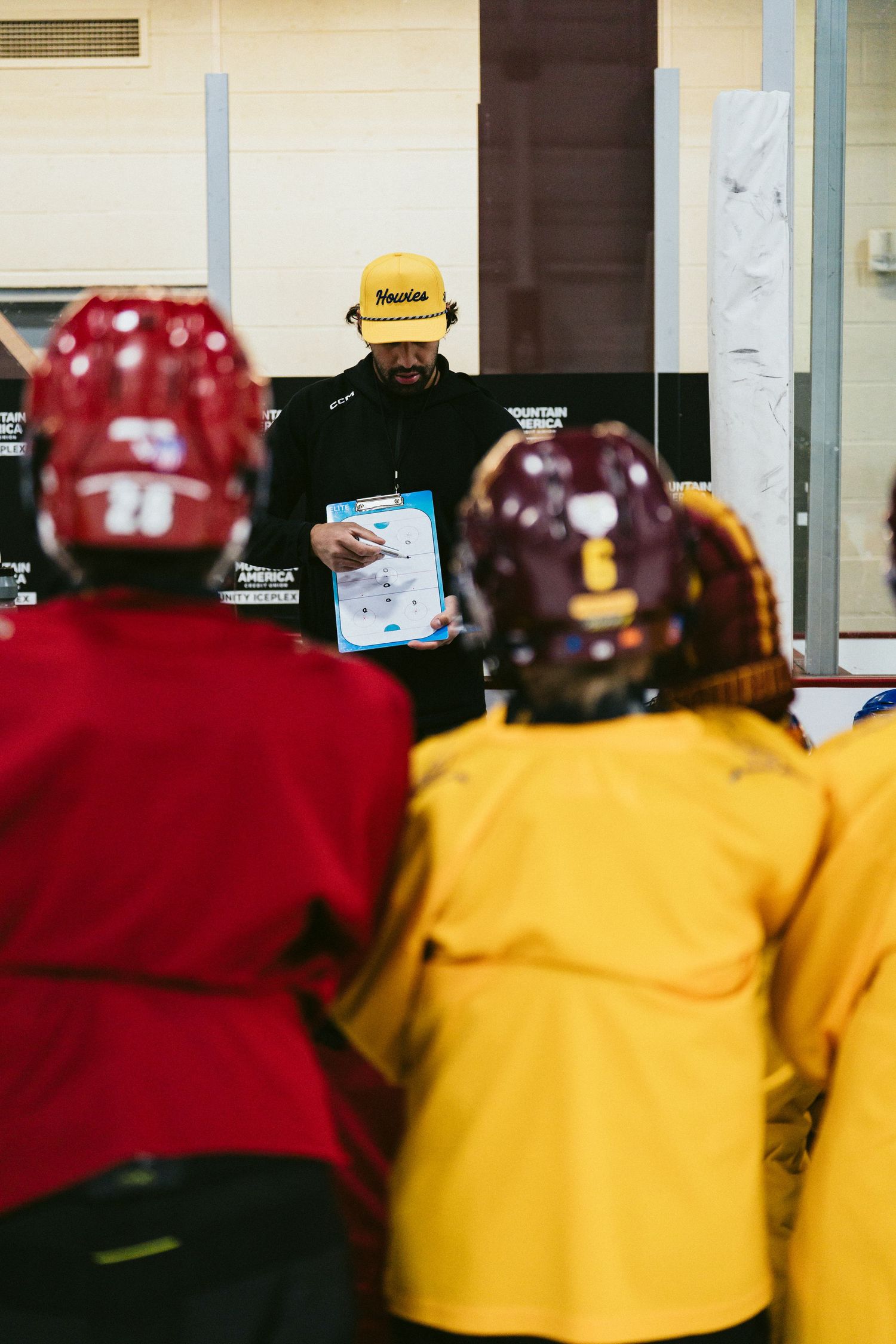 Hockey coach in yellow cap holds clipboard, giving instructions to youth players in red and yellow jerseys on ice rink.