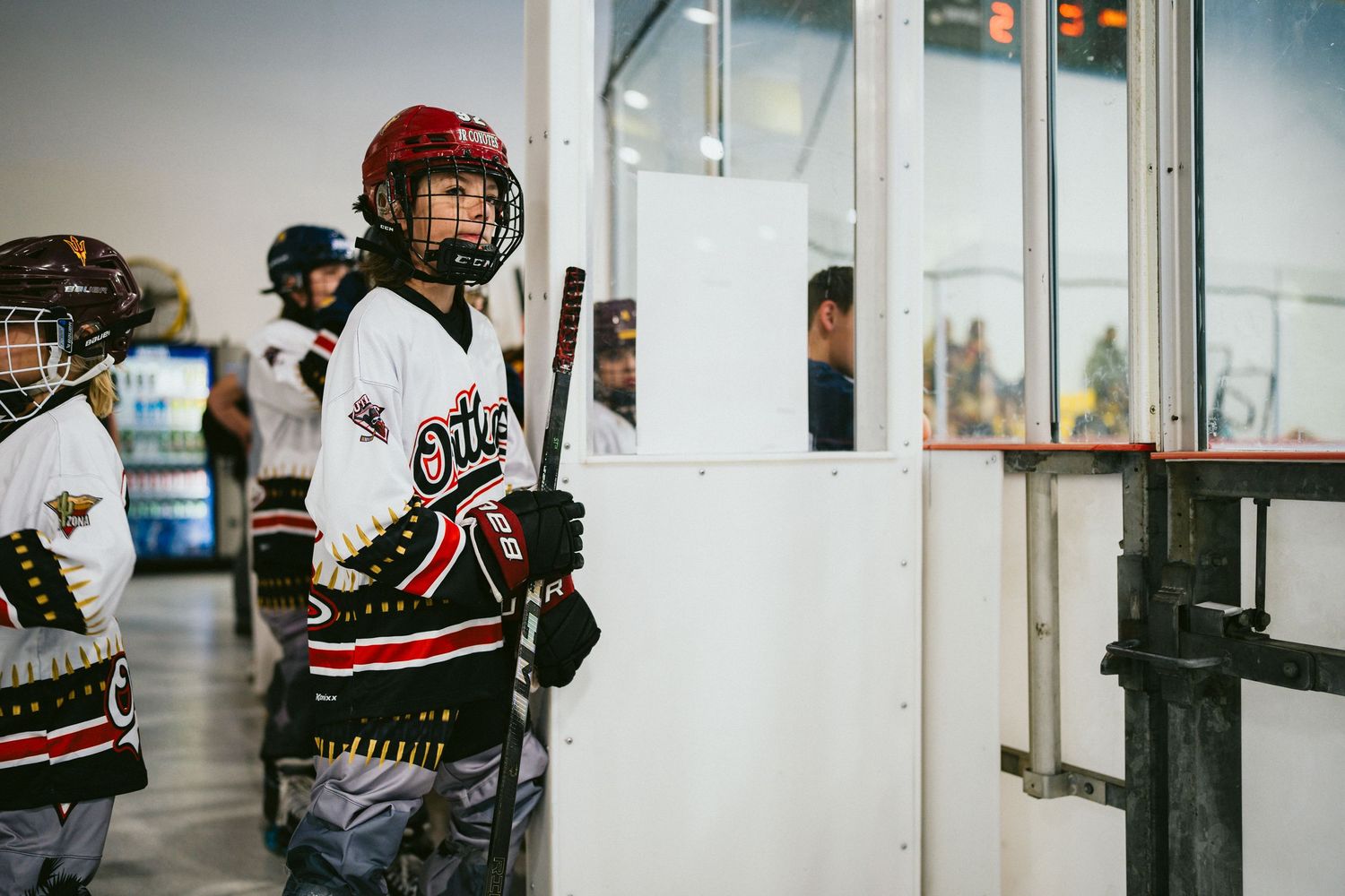 Young hockey player in Oilers jersey and red helmet waits by the bench door at an indoor roller hockey rink.