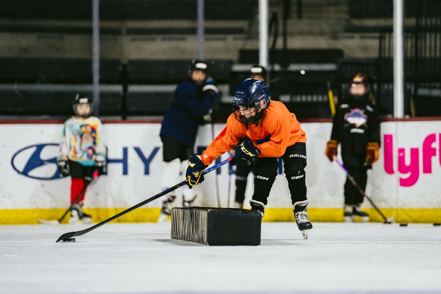 Young hockey player in orange jersey practices stickhandling with a training puck on an indoor rink.