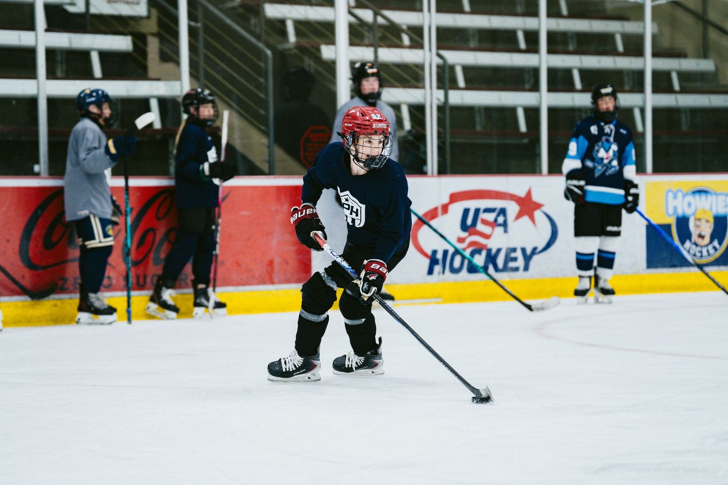 Young hockey player in red helmet skates with stick during USA Hockey practice at indoor rink.