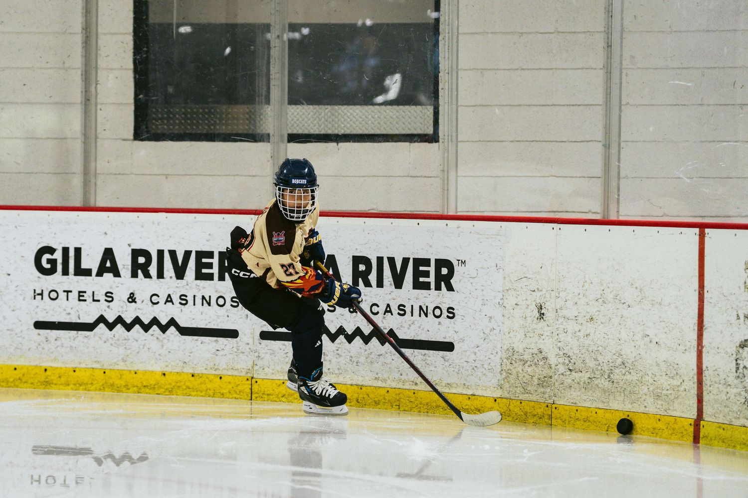 Young hockey player in black and gold uniform skating on ice rink with Gila River Hotels & Casinos boards.