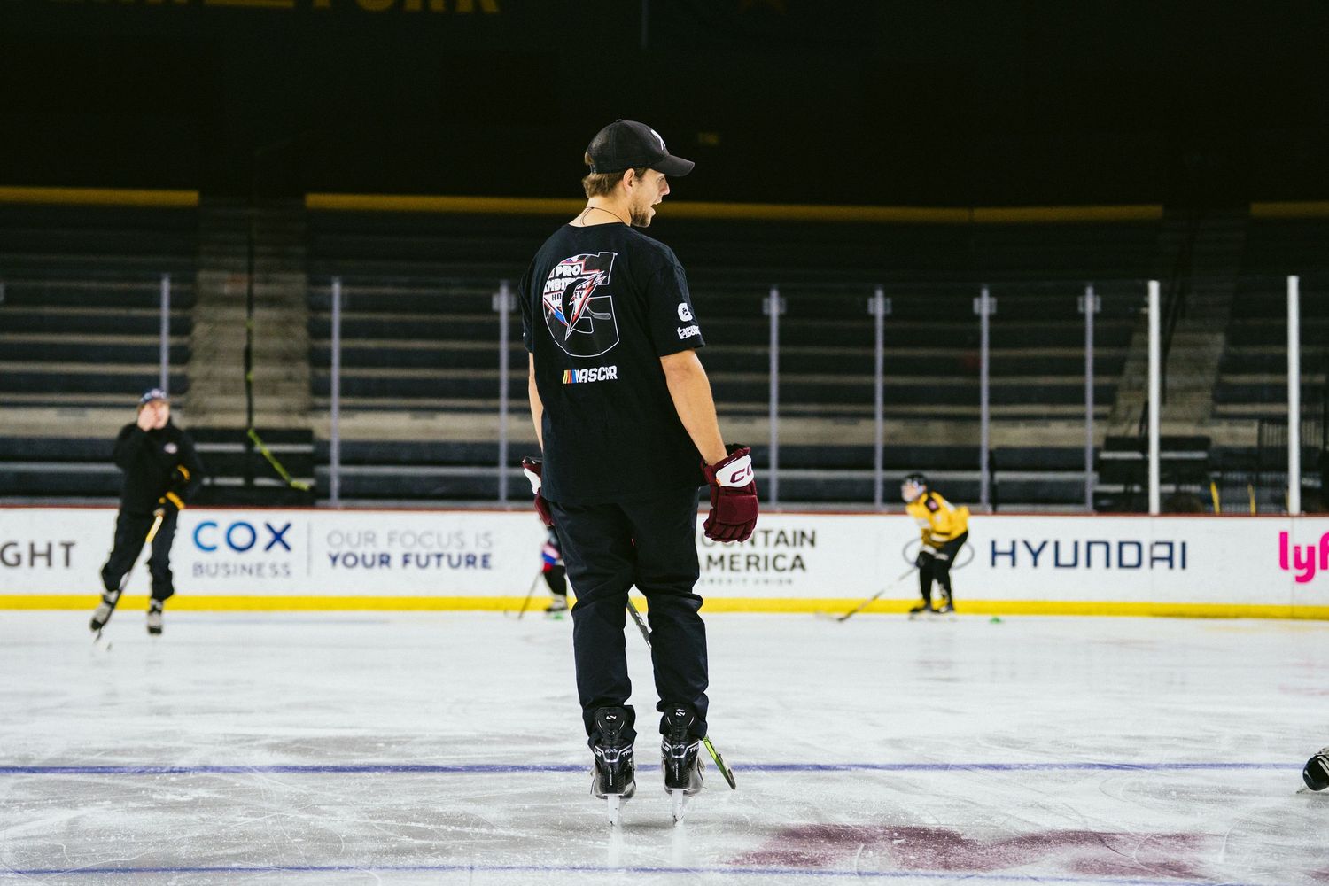 Hockey coach in black t-shirt stands on ice observing youth players during indoor rink practice.