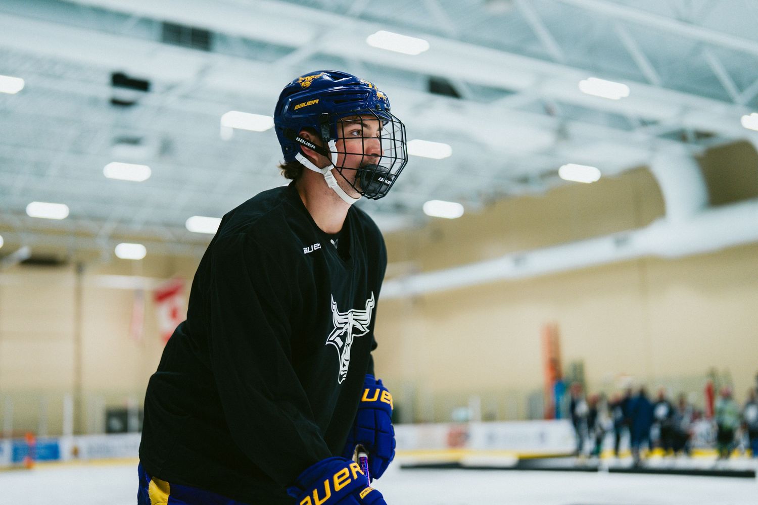 Ice hockey player wearing a blue Bauer helmet and team jersey skating during an indoor practice session at a hockey rink.