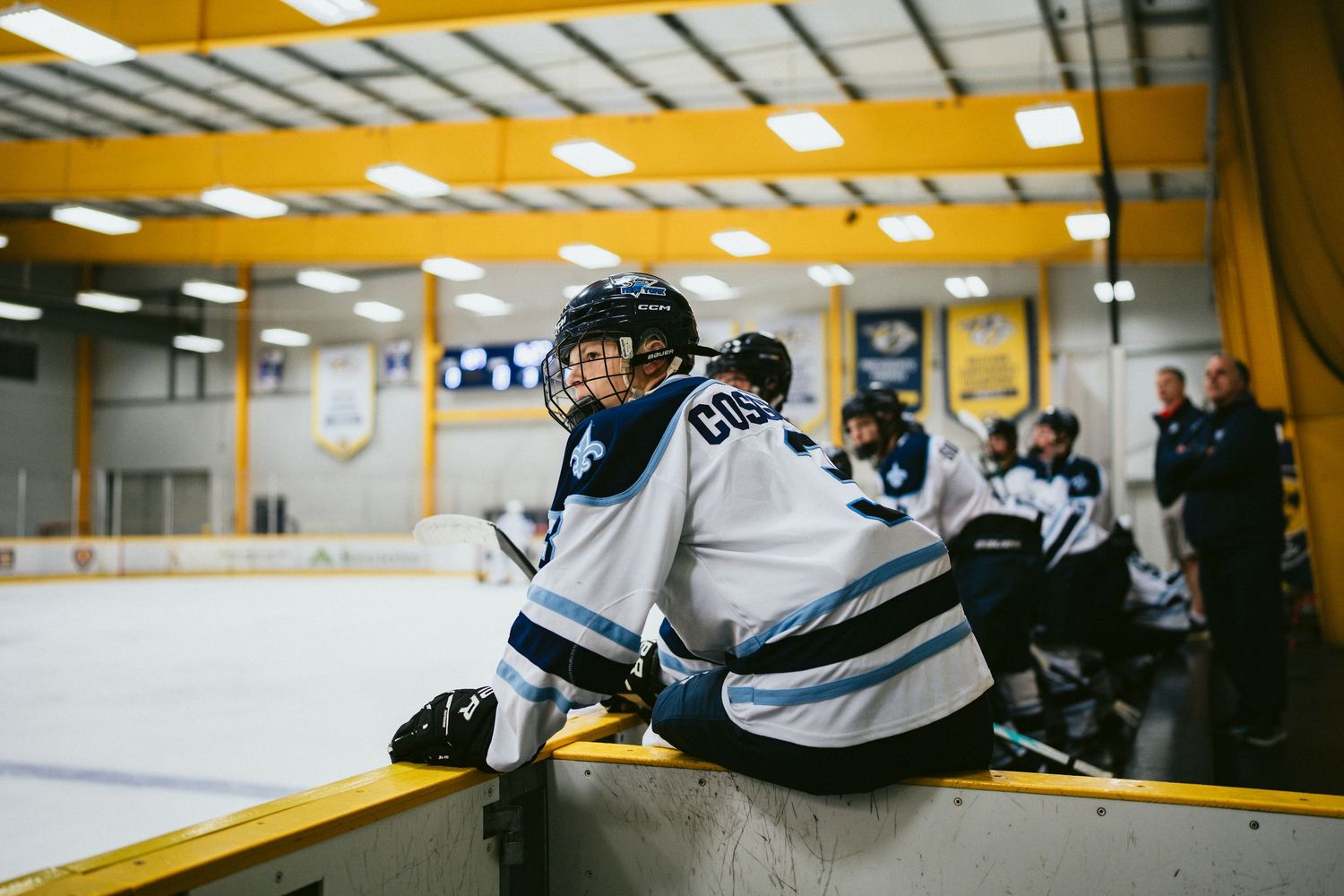 Young hockey player in blue and white jersey leans on boards during bench break at rink.