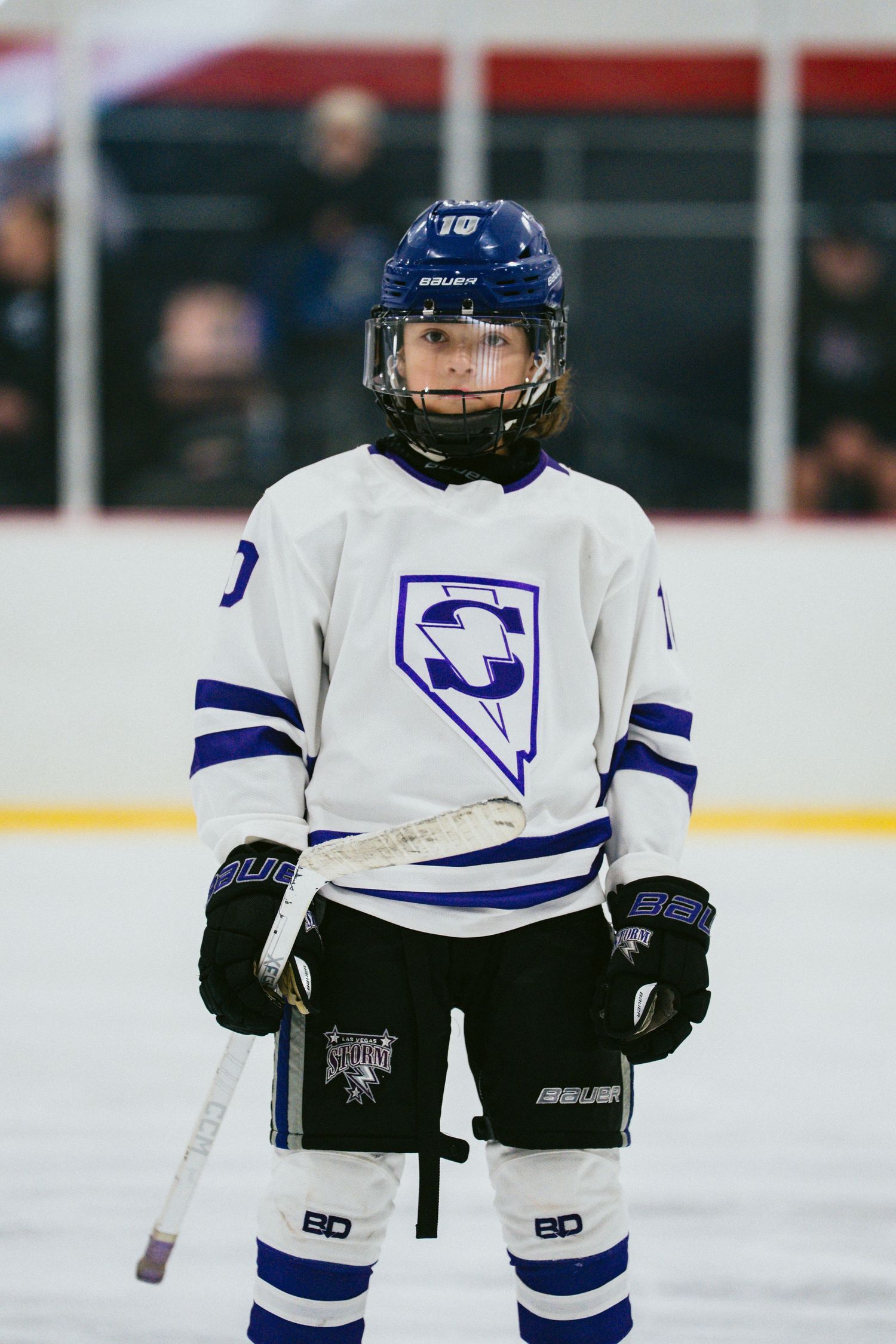 Youth hockey player in white jersey with blue stripes stands ready on ice.