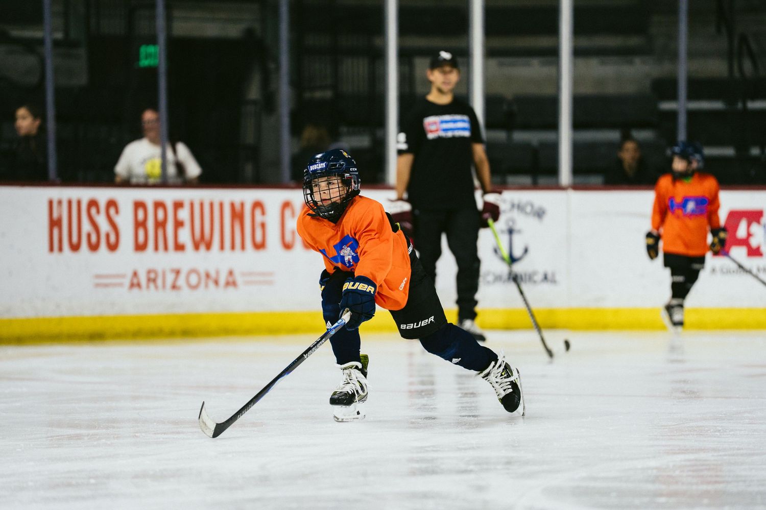 Young hockey player in orange jersey skating with puck at Huss Brewing Co Arizona ice rink.