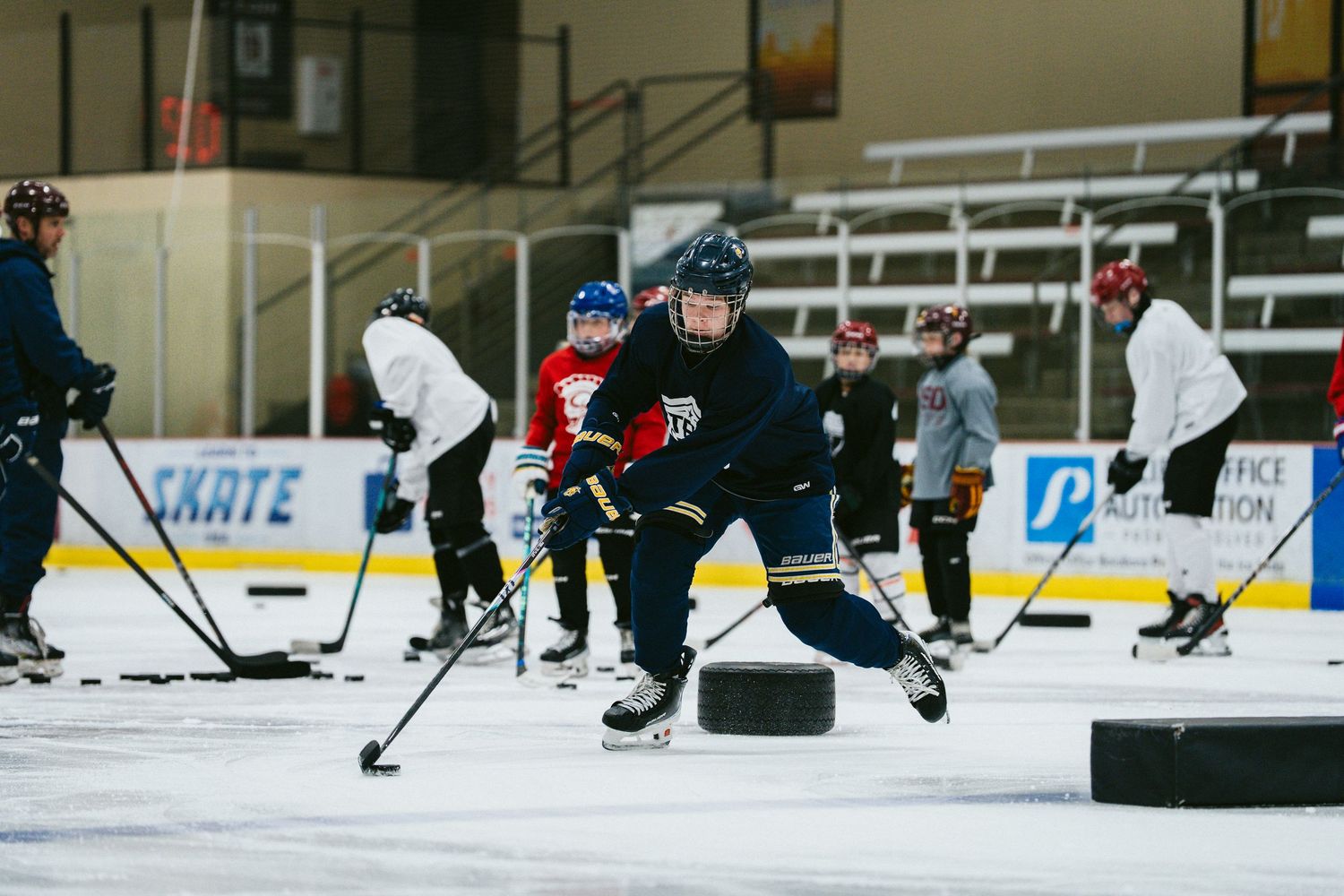 Young hockey player in navy jersey handles puck during ice hockey practice drill with teammates in background.