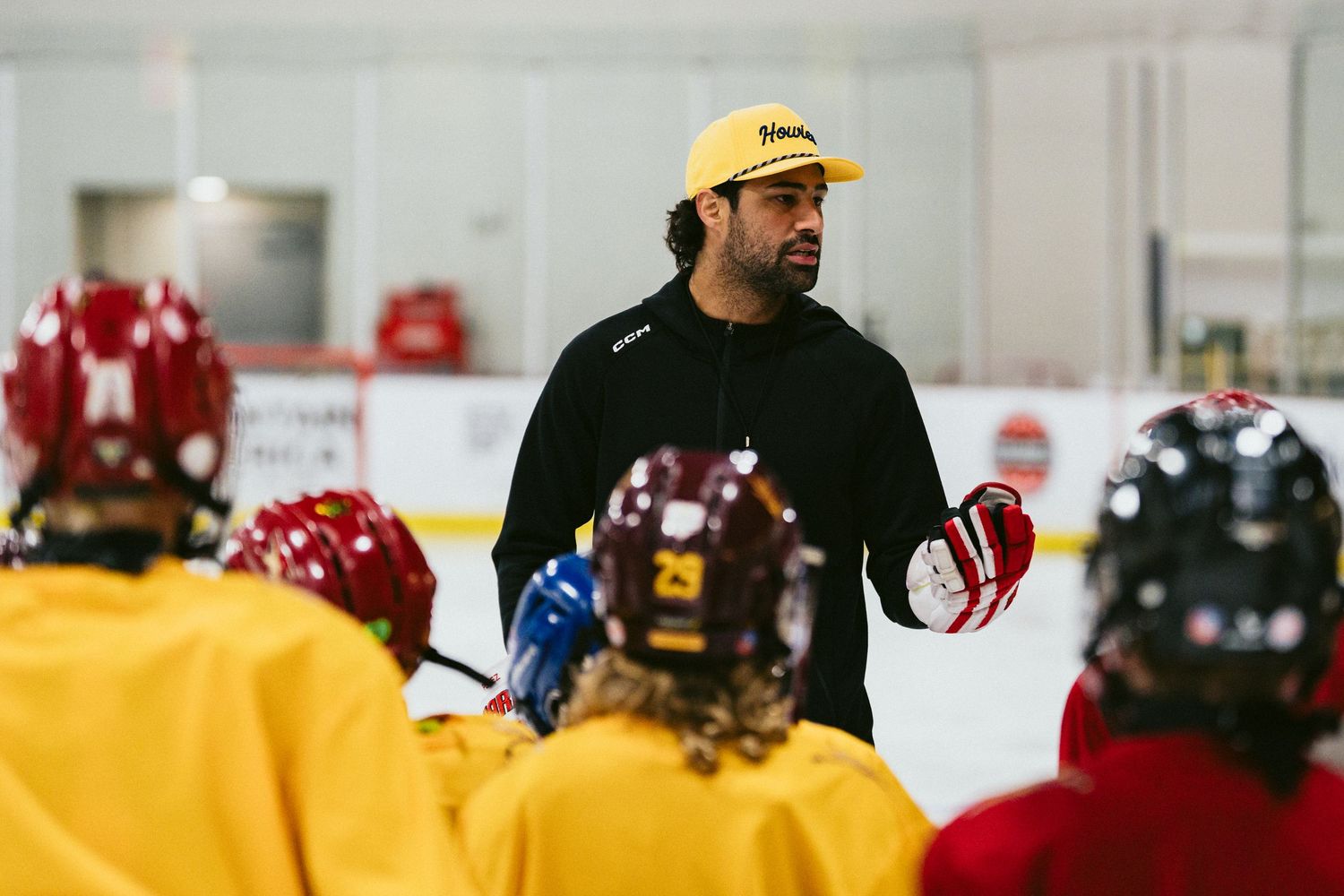 A hockey coach in a yellow cap addresses young players in yellow jerseys during an indoor ice rink practice.